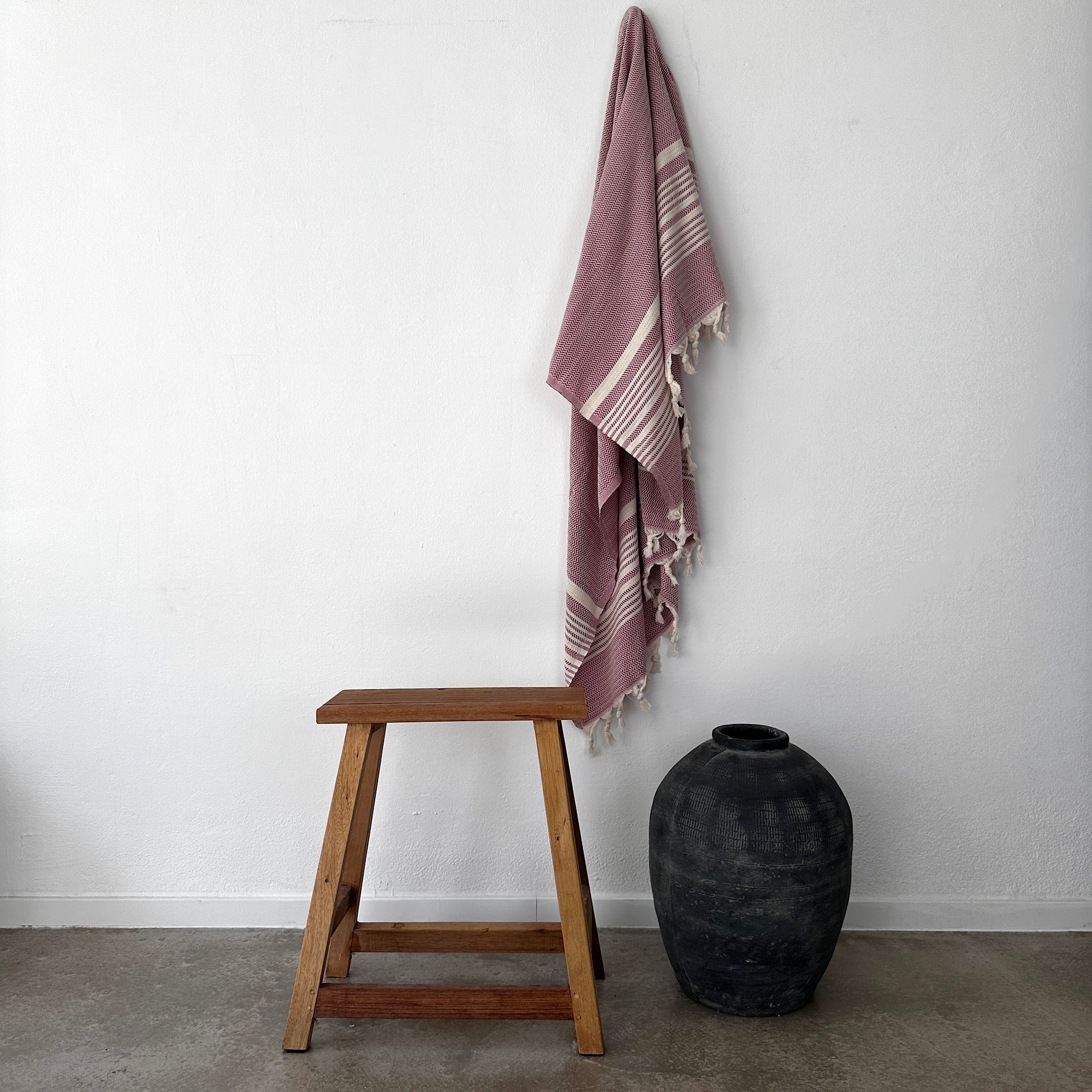 Ein Holzhocker steht neben einer großen schwarzen Vase auf dem Betonboden; ein auberginefarbenes Zickzack-Handtuch von LEY! Plaid-Handtuch von LEY! mit ausgefransten Rändern hängt an der weißen Wand über ihnen.