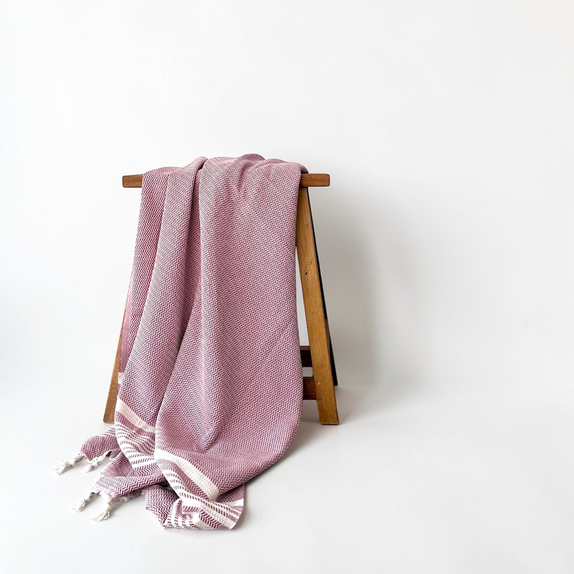 Das LEY! Plaid - Zickzack - Aubergine von LEY! mit Zickzack-Muster und Fransenkanten ist über einen Holzständer drapiert, der vor einem schlichten weißen Hintergrund steht.