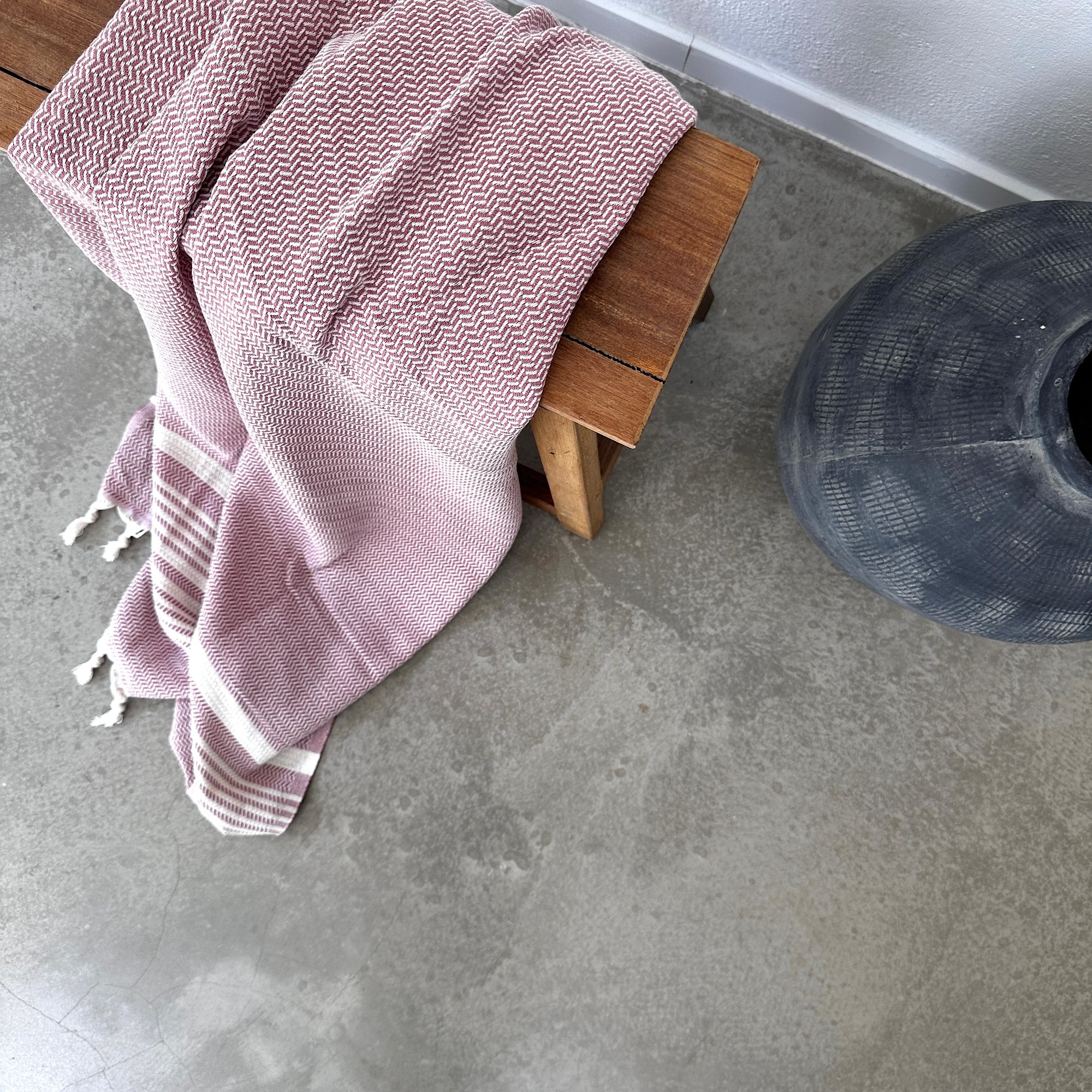 EIN LEY! Plaid - Zickzack - Aubergine ist über eine Holzbank neben einer großen schwarzen Vase auf einem glatten Betonboden drapiert - ein eleganter Look, der von LEY!.