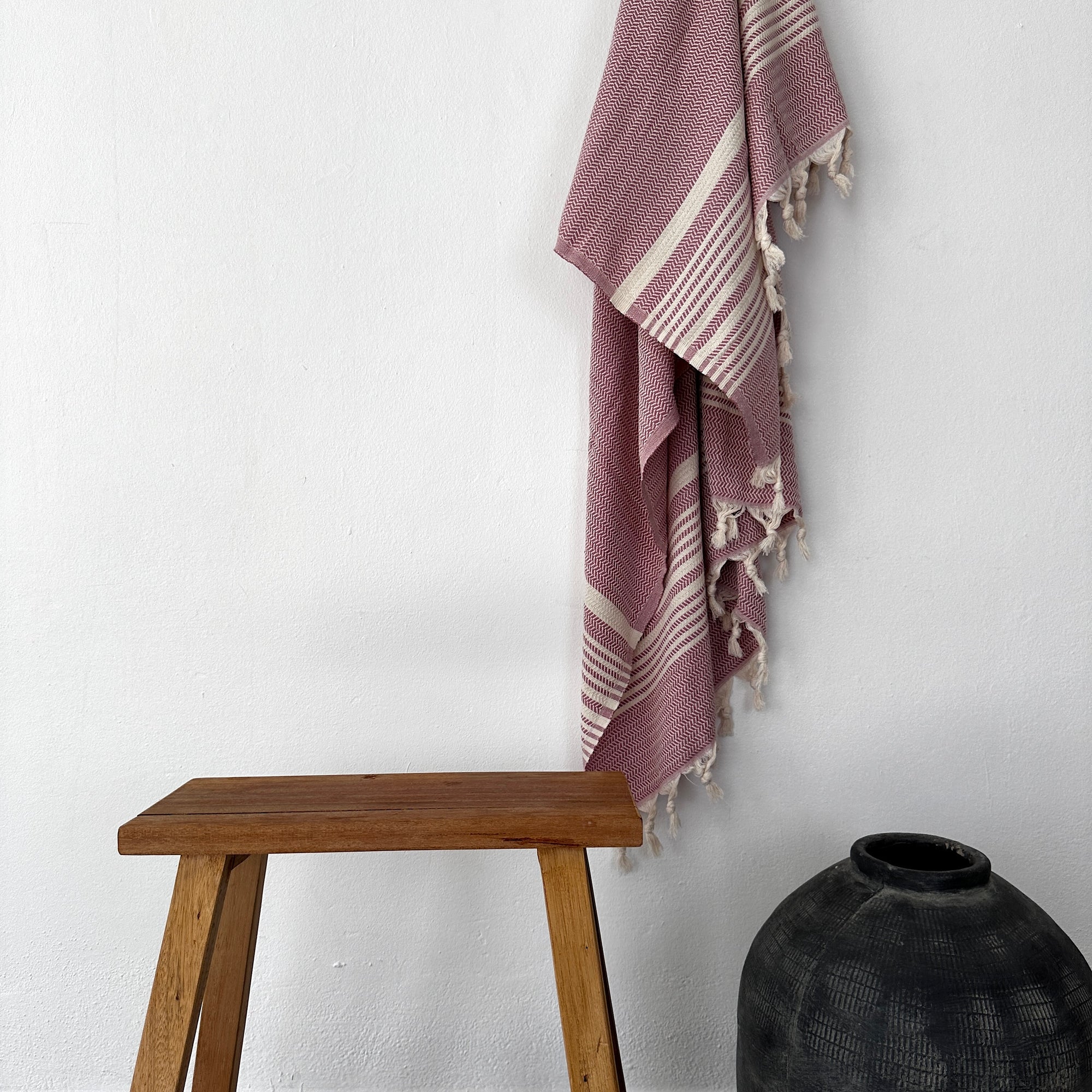 Ein Holzhocker steht neben einer dunklen Keramikvase, darüber hängt ein LEY! Plaid - Zickzack - Aubergine, das an der weißen Wand darüber hängt.