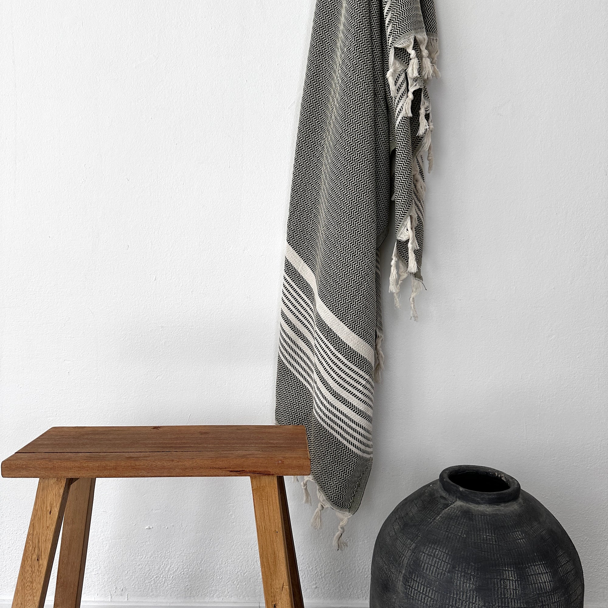 Ein Holzhocker steht neben einer strukturierten schwarzen Vase auf dem Boden, darüber hängt das LEY! Plaid - Zickzack - Olive von LEY! in Schwarz und Weiß an der weißen Wand über ihnen hängt.