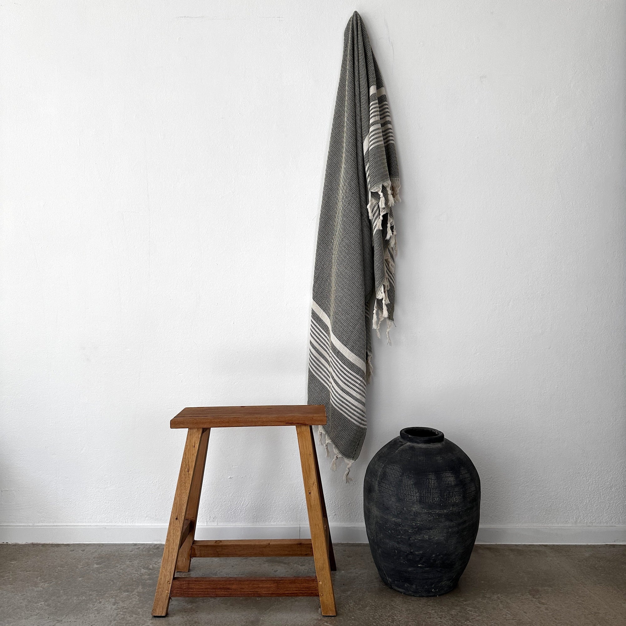 Das LEY! Plaid - Zickzack - Olive von LEY! ist an einer weißen Wand drapiert, daneben stehen ein Holzhocker und eine große schwarze Keramikvase, alles auf einem Betonboden in einem minimalistischen Raum.