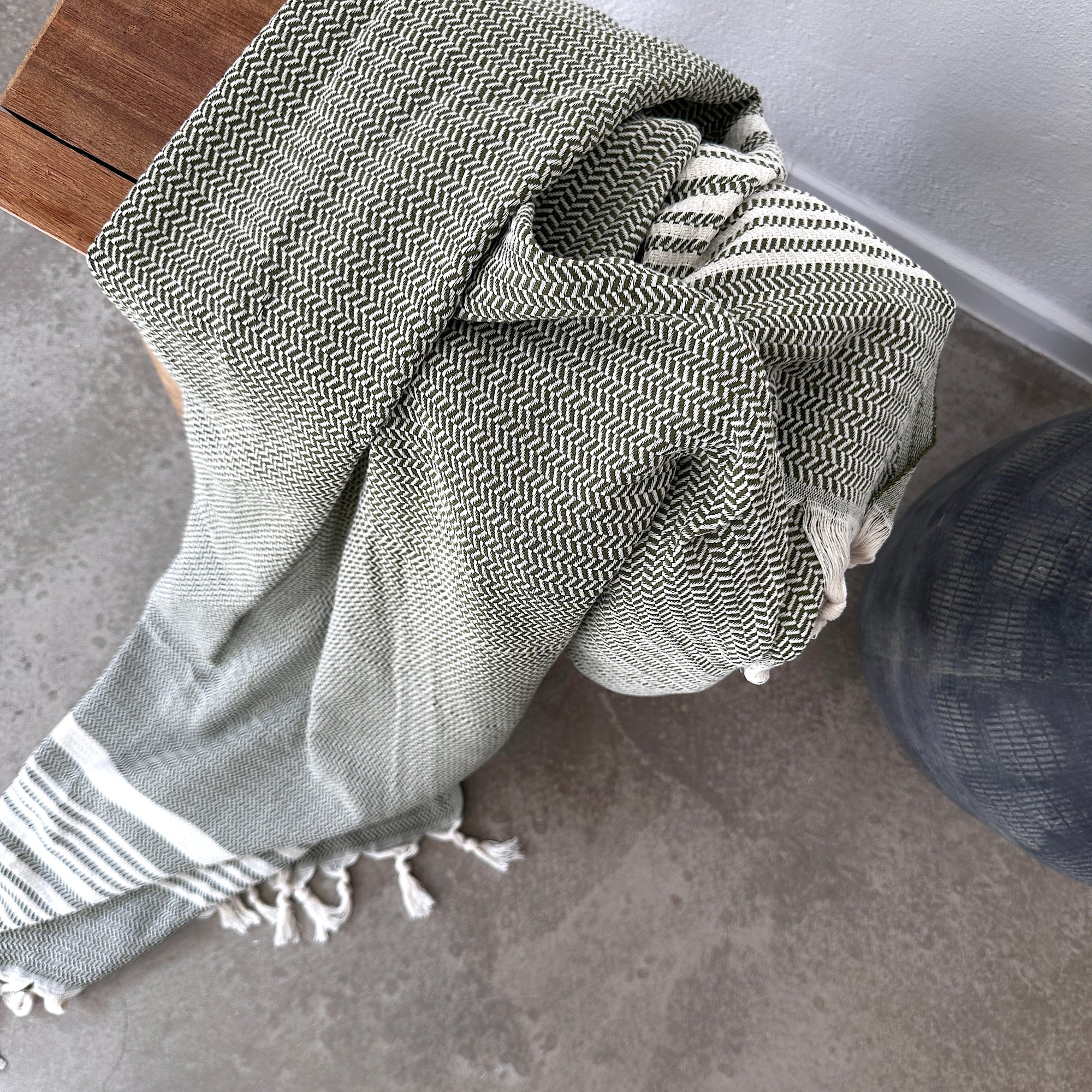 Die LEY! Plaid - Zickzack - Olive, ein grün-weiß strukturierter Überwurf mit Fransen von LEY!, ist über einer Holzbank auf grauem Betonboden neben einem dunklen, runden Objekt drapiert.