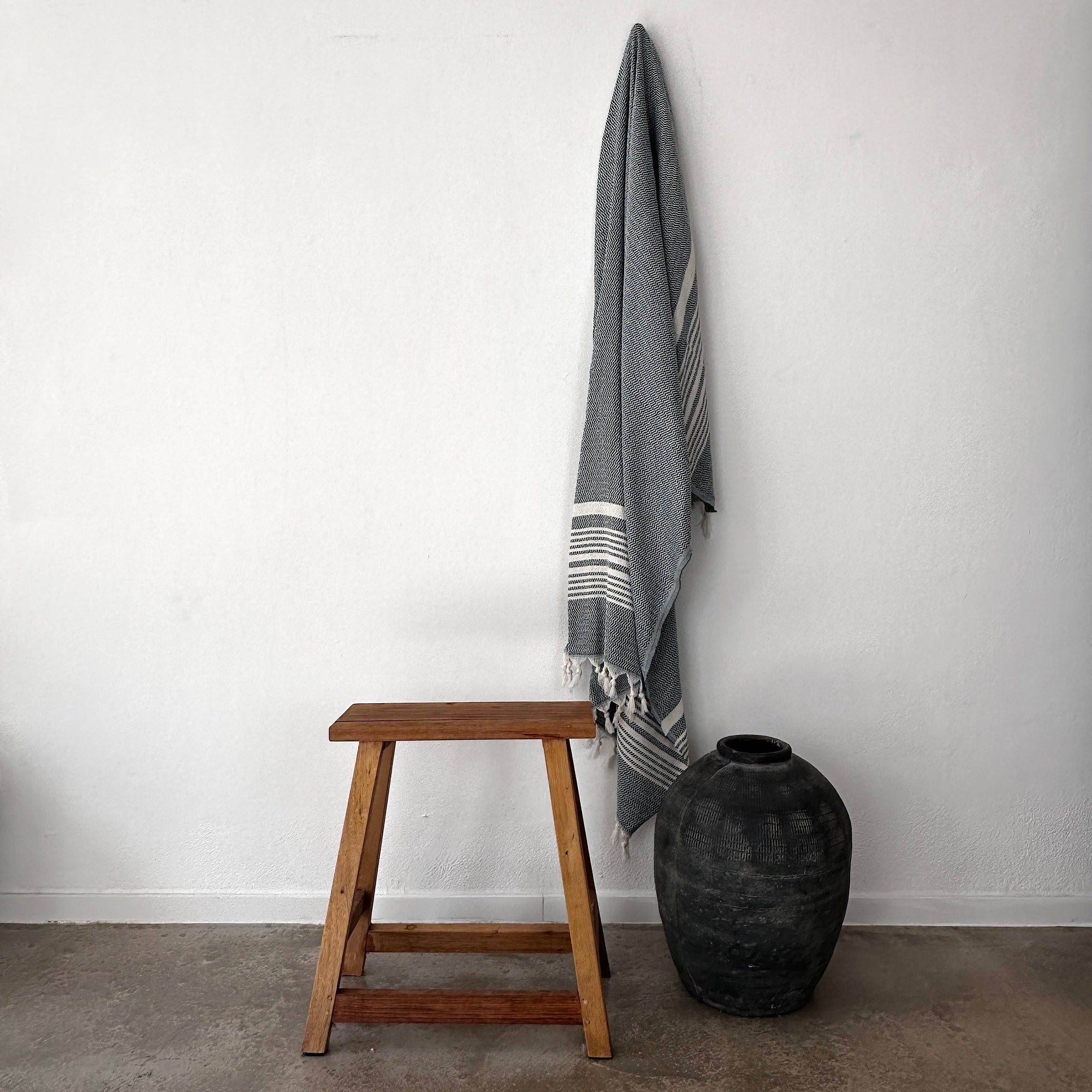 Eine minimalistische Szene mit einem Holzhocker, einer großen schwarzen Keramikvase und einer LEY! Plaid - Zickzack - Petroldecke an einer weißen Wand in einem einfachen, modernen Interieur.
