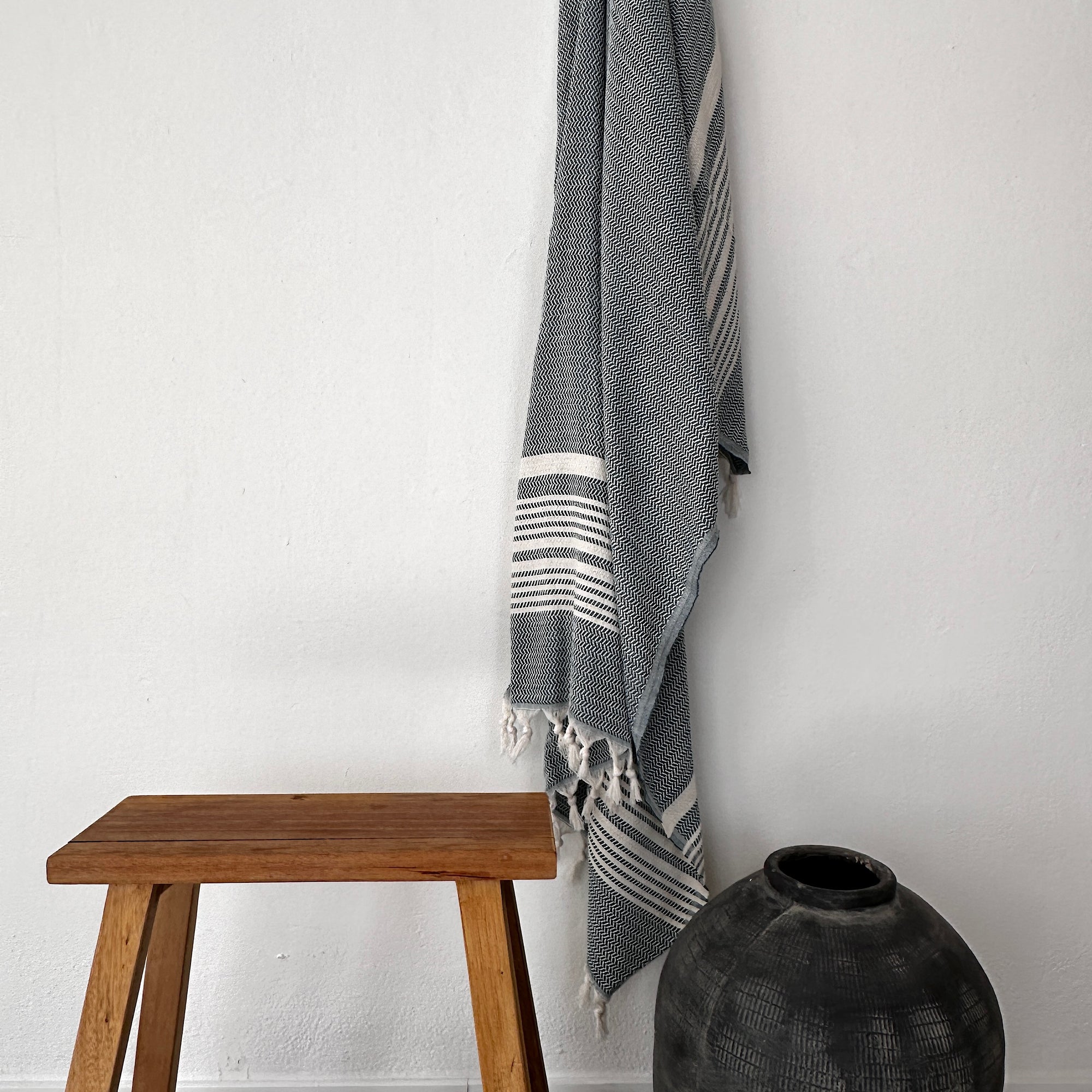 EIN LEY! Plaid - Zickzack - Petrol von LEY! hängt an einer weißen Wand über einer einfachen Holzbank. In der Nähe steht eine dunkle, runde Keramikvase auf dem Boden und vervollständigt das minimalistische, moderne Dekor.