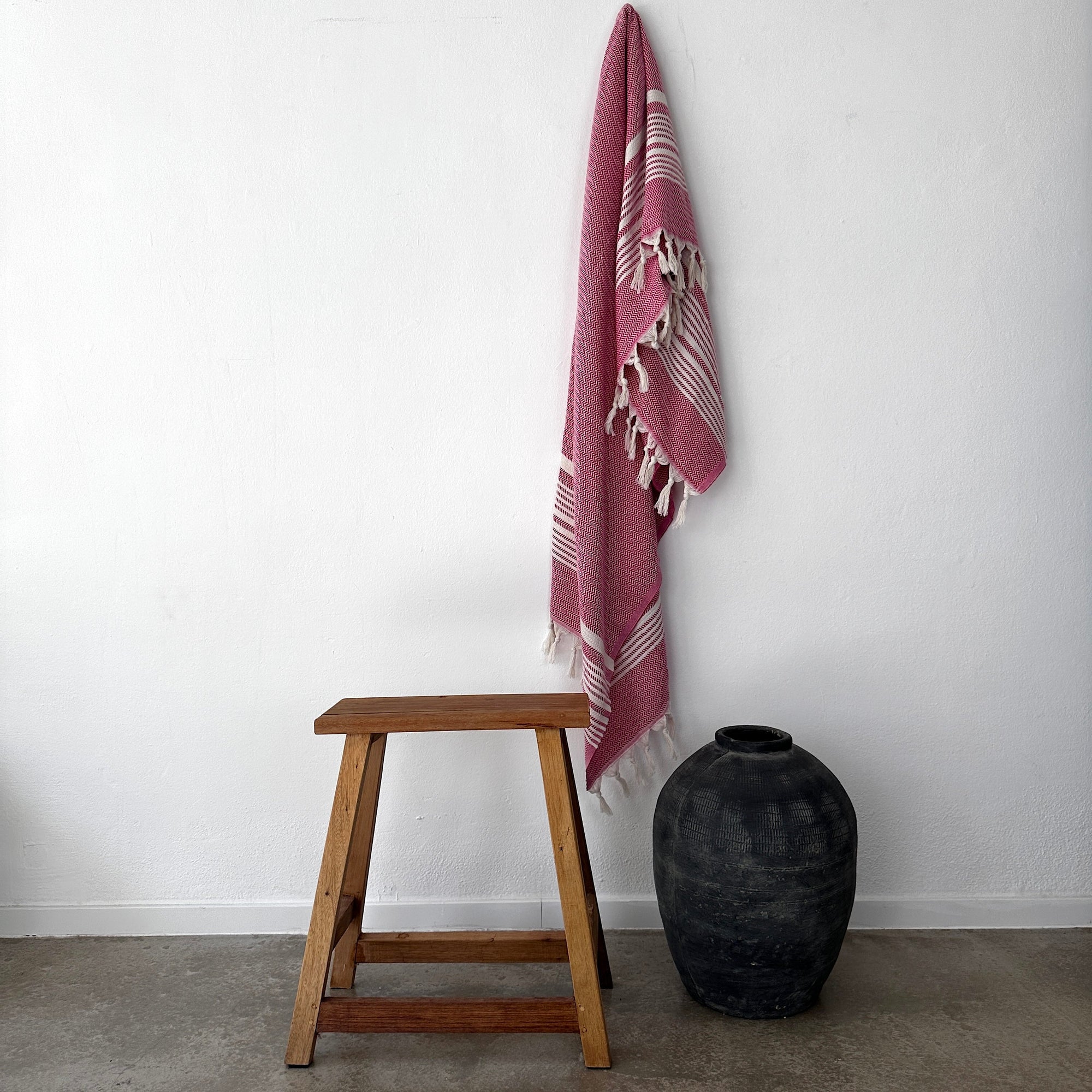 Ein Holzhocker steht neben einer großen schwarzen Keramikvase auf einem Betonboden, darüber hängt die LEY! Plaid - Zickzack - Pink von LEY! hängt an der weißen Wand über ihnen.