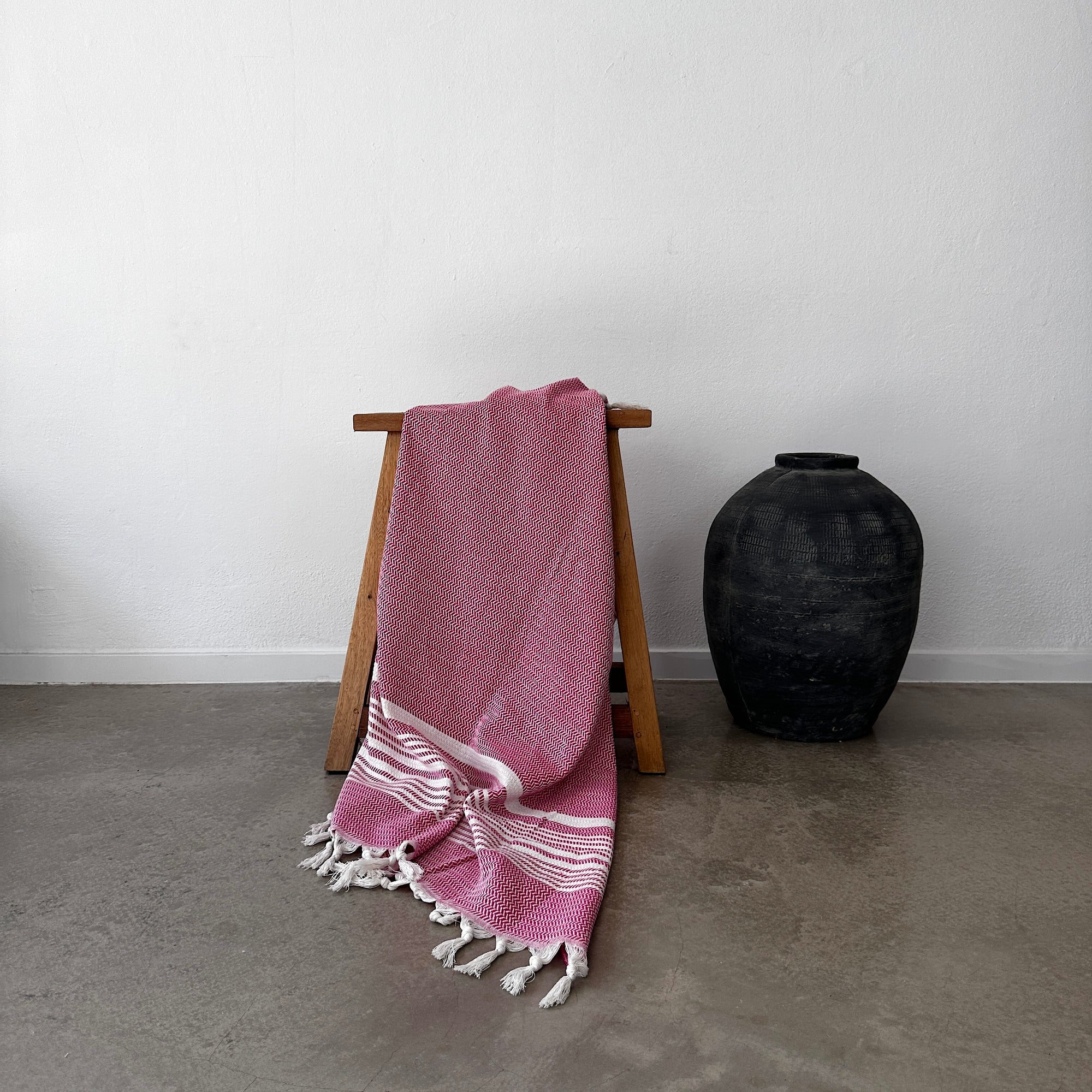 Das LEY! Plaid - Zickzack - Pink von LEY! mit weißen Fransen ist über einen Holzständer neben einer schwarzen Keramikvase auf einem grauen Betonboden vor einer schlichten weißen Wand drapiert.
