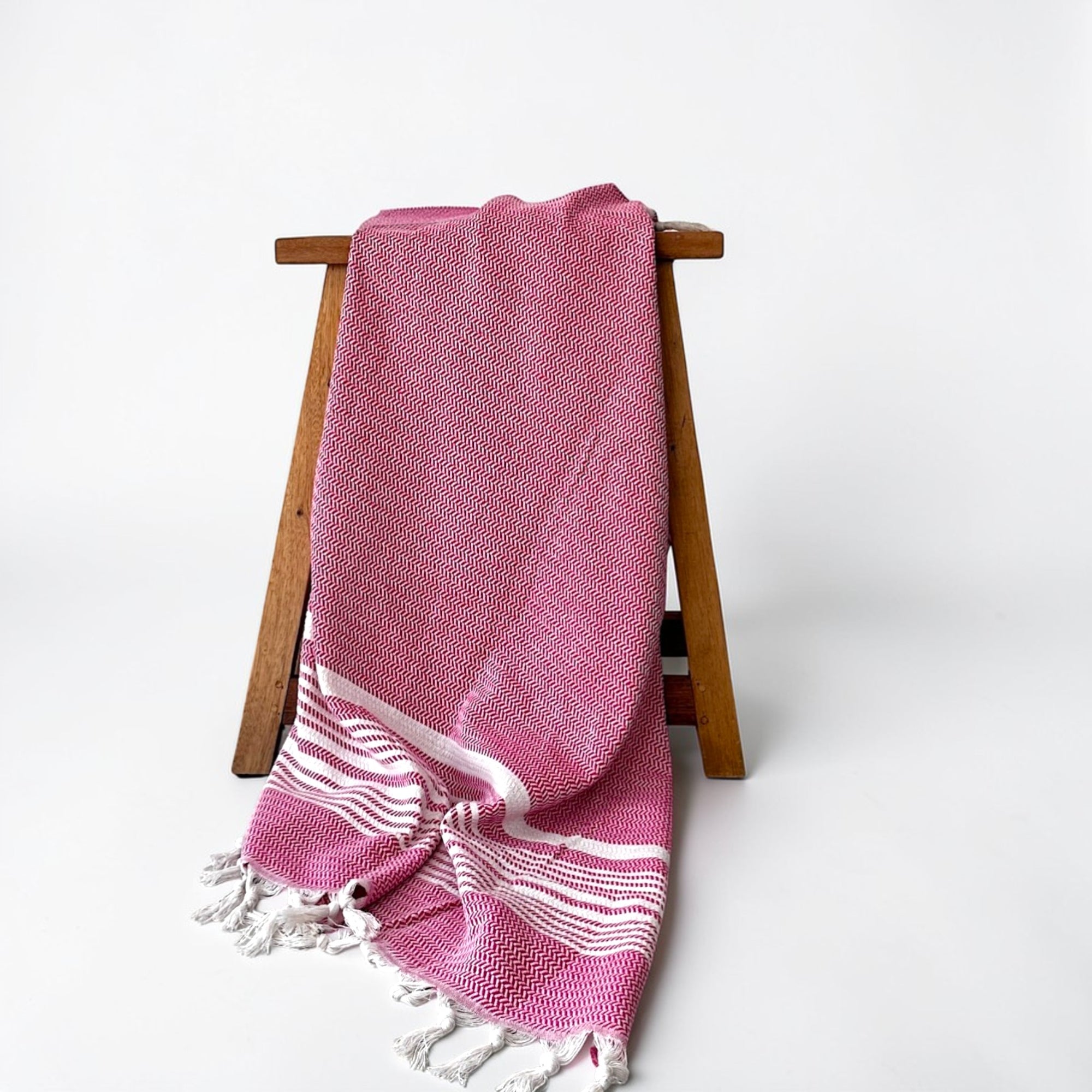 Das LEY! Plaid - Zickzack - Pink von LEY! mit pinkfarbenen und weißen Zickzack-Webdetails und geflochtenen Rändern wird über einen Holzhocker vor einem schlichten weißen Hintergrund drapiert.