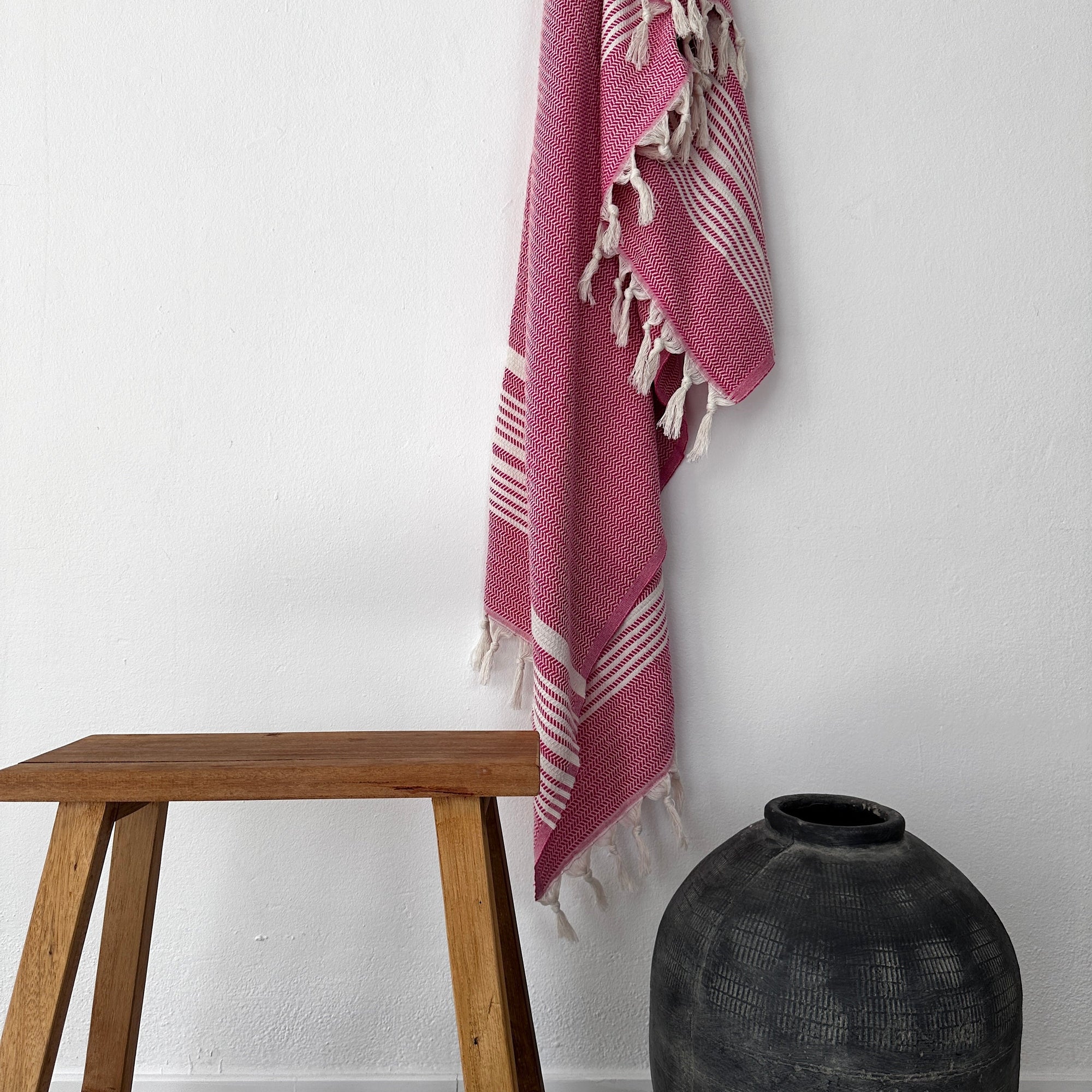 Ein rosa-weißes LEY! Plaid - Zickzack hängt mit Quasten an einer weißen Wand über einem Holzhocker, neben einer dunkelgrauen Keramikvase - ein einladendes Stück von LEY!.
