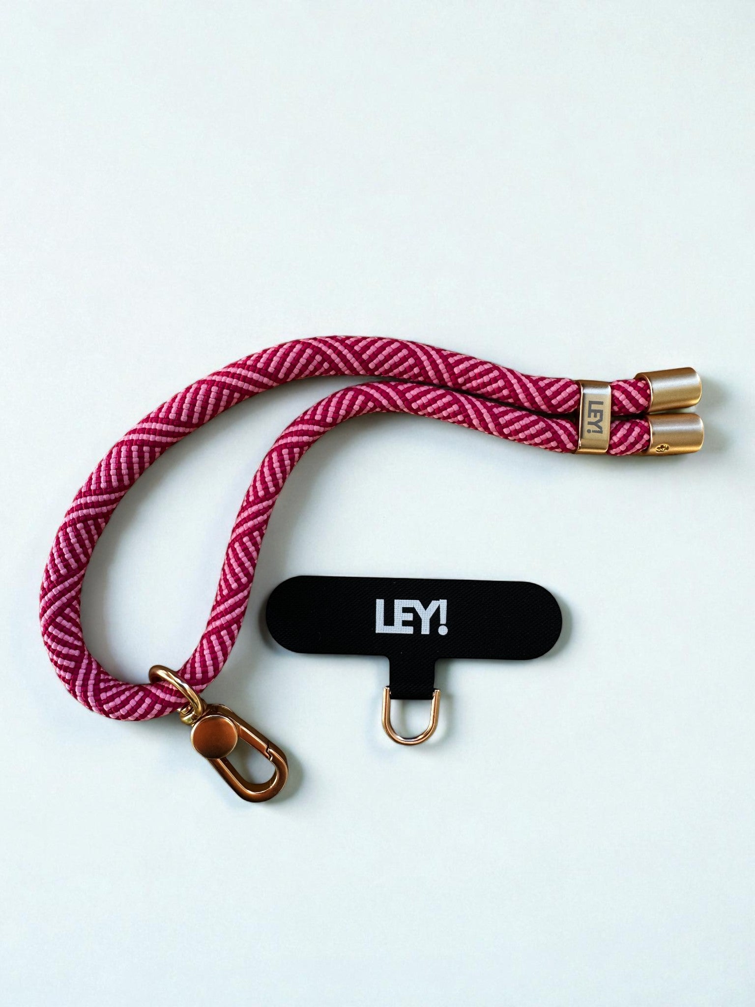 Der LEY! Schlüsselanhänger ist ein rosa-weiß geflochtenes Schlüsselband mit goldenen Metallenden, einem Clip, einem schwarzen "LEY!"-Schriftzug in weißer Schrift und einem kleinen goldenen Haken - stylisch und praktisch, um dein Handy immer griffbereit zu haben.