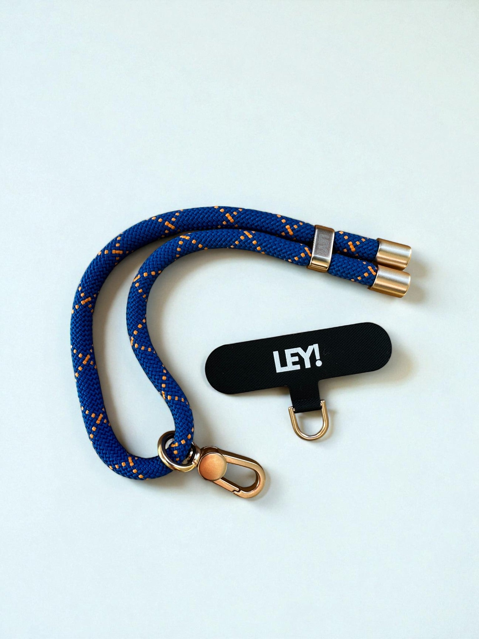 Ein blauer gewebter Schlüsselanhänger von LEY! mit orangefarbenen Akzenten, goldenen Metallenden und einer goldenen Schließe liegt neben einem schwarzen "LEY!"-Anhänger in Weiß und einem kleinen Ring - ideal, um dein Handy stets griffbereit in trendigen Farben zu halten.