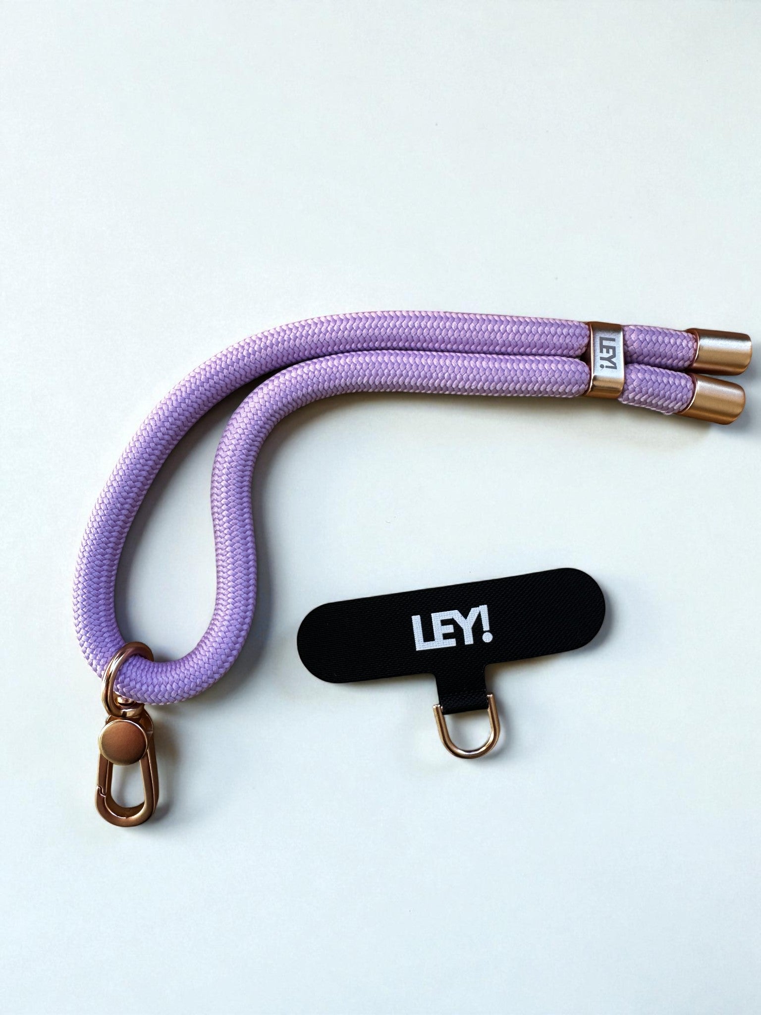 Ein lavendelfarbener Schlüsselanhänger Handyband von LEY! mit goldenen Metallenden und Schließe liegt neben einem schwarzen LEY!-Anhänger und Goldring. Halte dein Handy stets griffbereit - alles auf weißem Hintergrund.