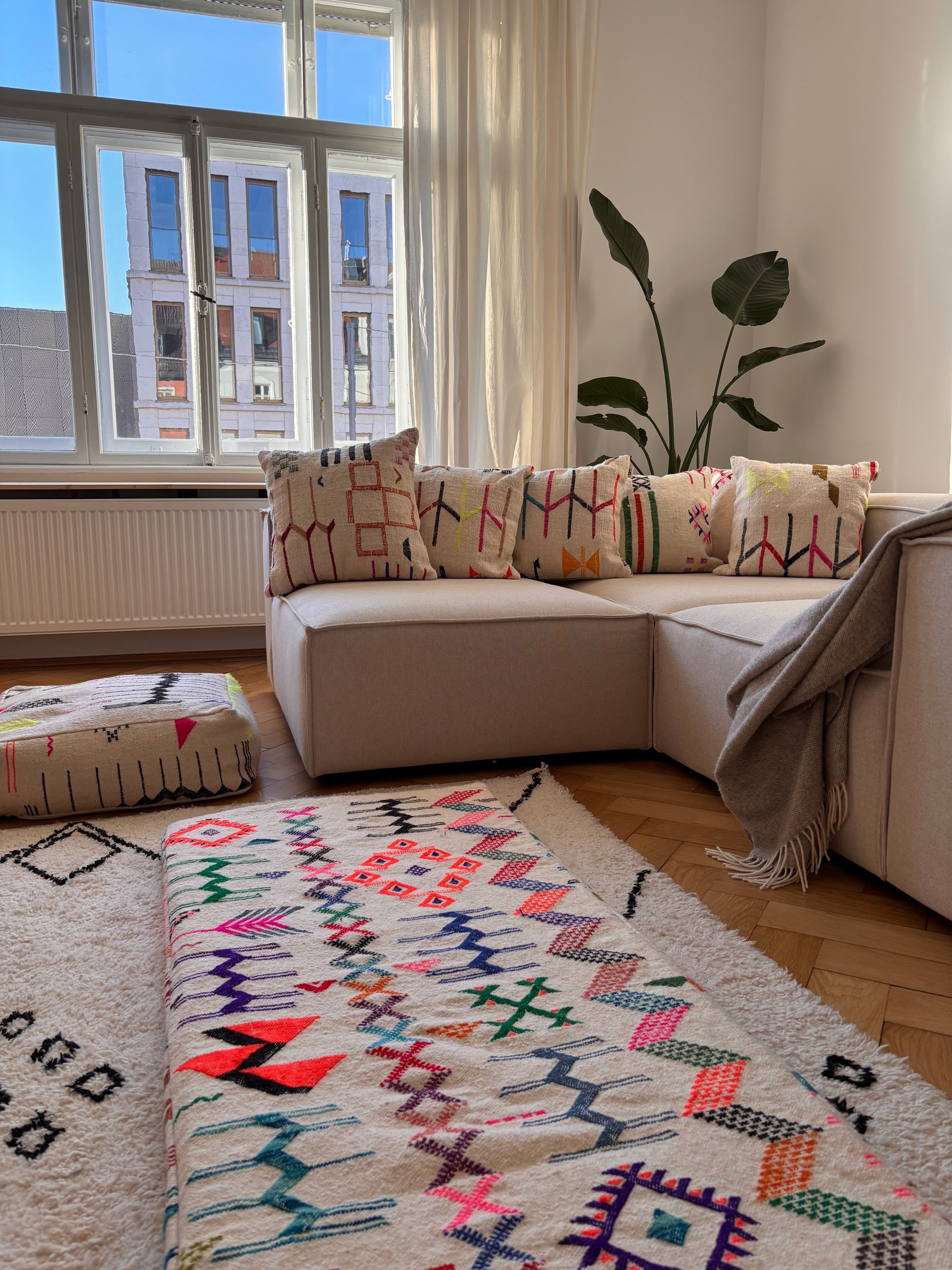 Ein gemütliches Wohnzimmer mit einem beigen Sektionssofa, gemusterten Kissen, einer grauen Decke, einer großen Blattpflanze und bunten geometrischen Teppichen auf einem Holzboden. Durch ein großes Fenster strömt Sonnenlicht herein.