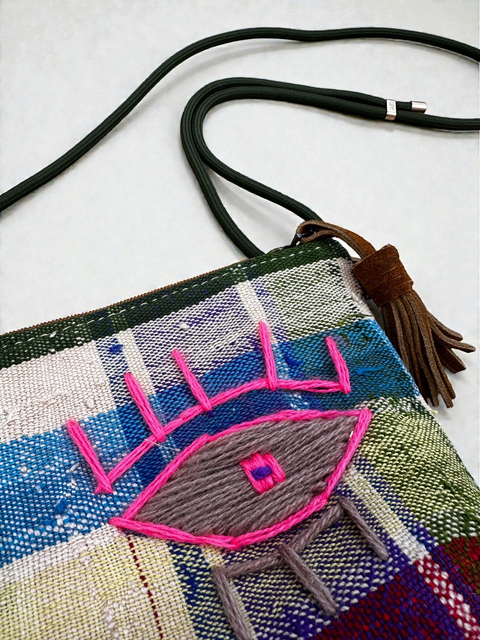 Die LEY! Tasche - OCULIA CLUTCH von LEY! ist eine handgefertigte Umhängetasche mit grünem Riemen, braunem Tassel und gesticktem Auge mit pinken Details auf blau-grün-weiß kariertem Hintergrund.