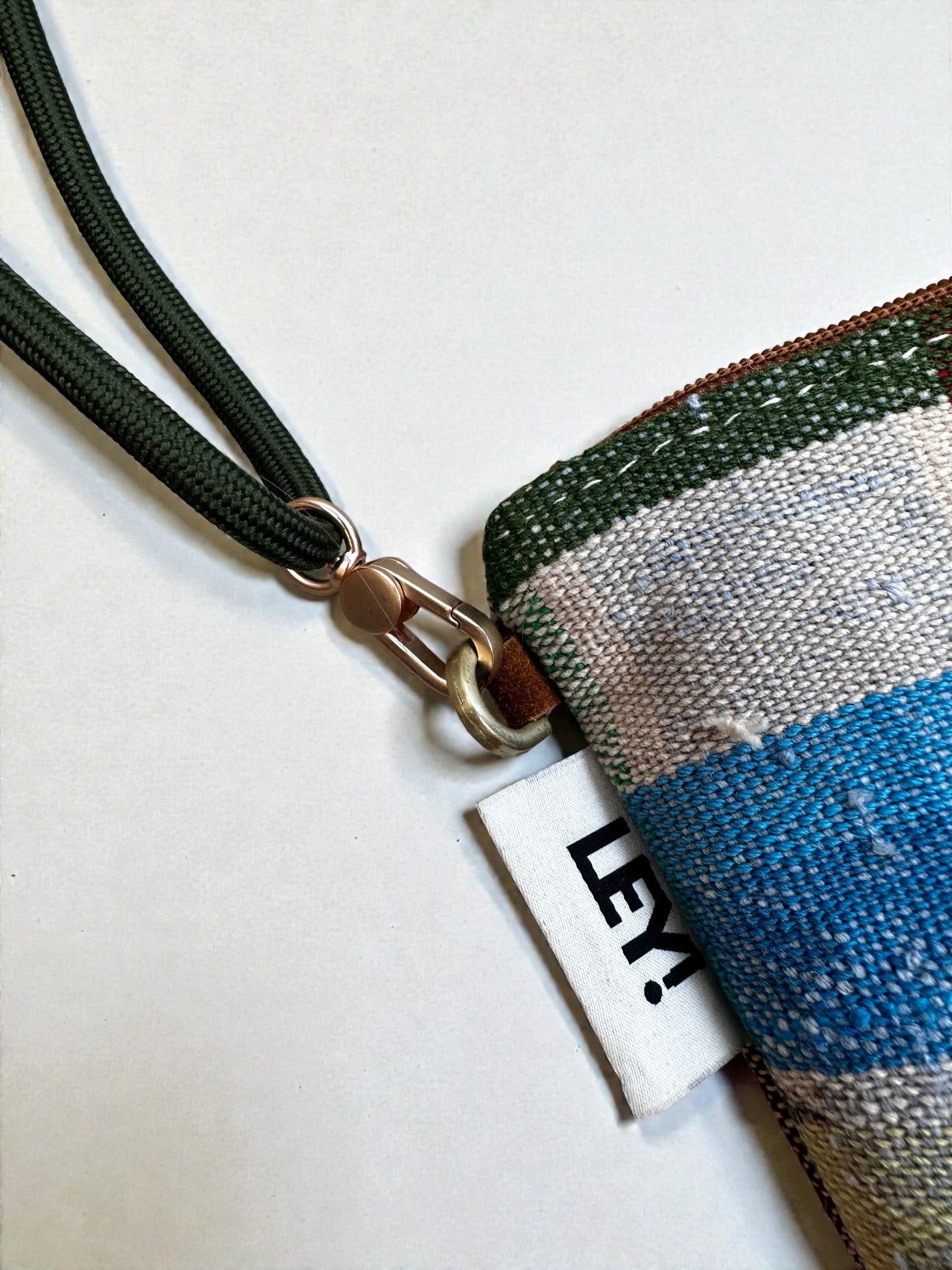 Eine Nahaufnahme der LEY! Tasche - OCULIA CLUTCH von LEY!, einer gewebten Tasche mit grünen, blauen und beigen Streifen an einem grünen Kordel-Schlüsselanhänger, mit urbanem Design und einem weißen Stoffanhänger mit der Aufschrift "LEY!".