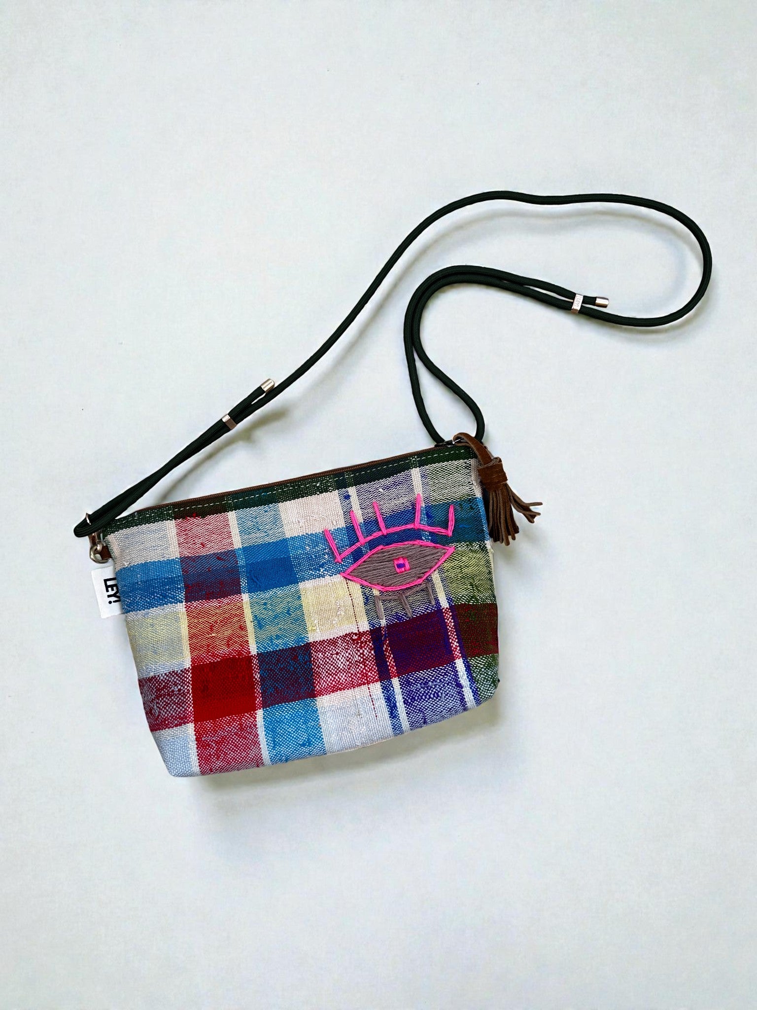 Die LEY! Tasche - OCULIA CLUTCH von LEY! ist eine kleine, handgefertigte Umhängetasche mit mehrfarbigem Karo, schwarzem Riemen und rosa Augenstickerei auf der Vorderseite. Die kompakte Tasche ist vor einem schlichten weißen Hintergrund abgebildet.