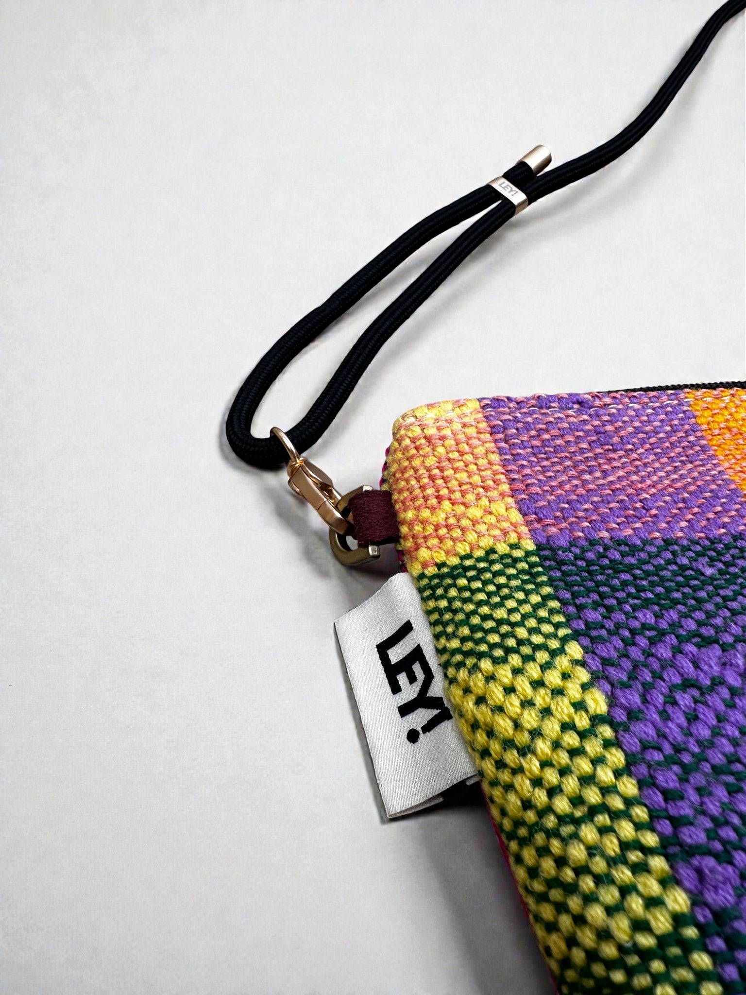 Eine Nahaufnahme der LEY! Tasche - OCULIA CLUTCH von LEY!, einer farbenfrohen, gewebten Clutch mit schwarzem Handgelenkband, weißem "LEY!"-Anhänger und einem leuchtend gelben, grünen, lila und orangefarbenen Karomuster auf hellem Grund.