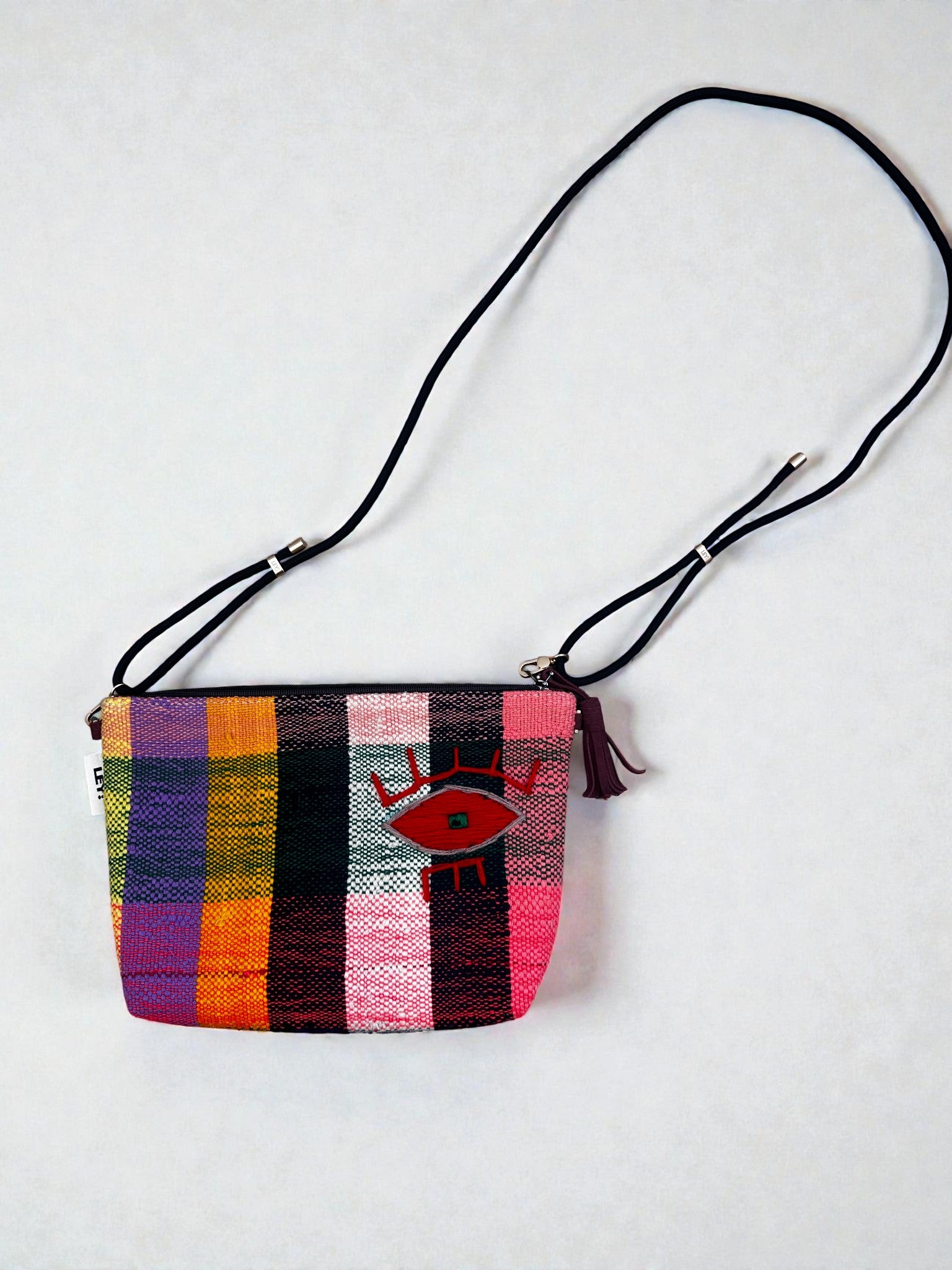 Die LEY! Tasche - OCULIA CLUTCH von LEY! ist eine farbenfrohe, handgefertigte Umhängetasche mit schwarzem, verstellbarem Riemen, Karomuster in Gelb-, Orange-, Grün-, Pink- und Lila-Tönen und gesticktem Augenmotiv.