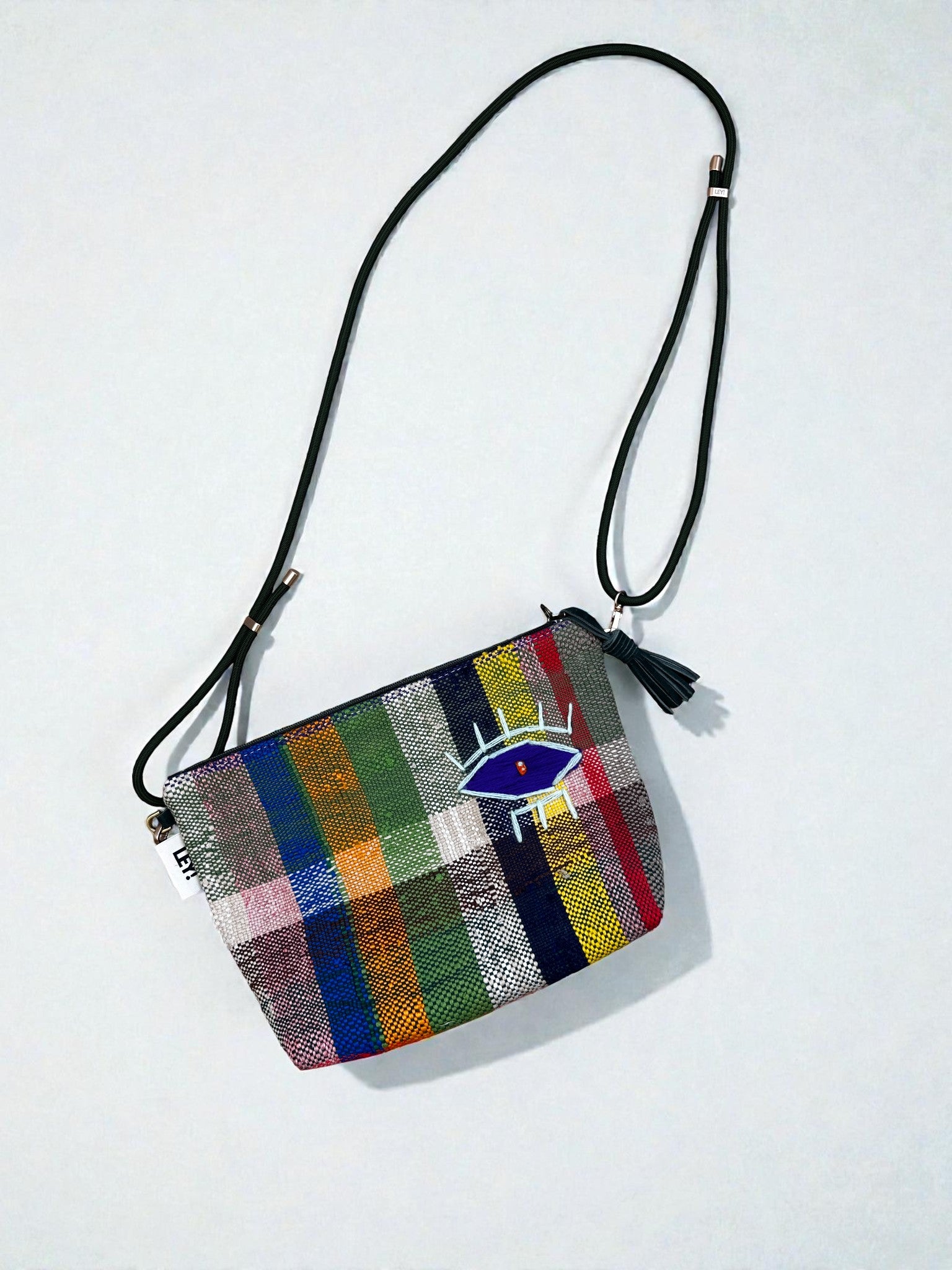 Die LEY! Tasche - OCULIA CLUTCH von LEY! ist eine kleine handgefertigte Crossbody-Tasche mit einem langen schwarzen Riemen, einem bunten Karomuster, einem gestickten blauen Auge auf der Vorderseite und einer schwarzen Quaste am Reißverschluss.