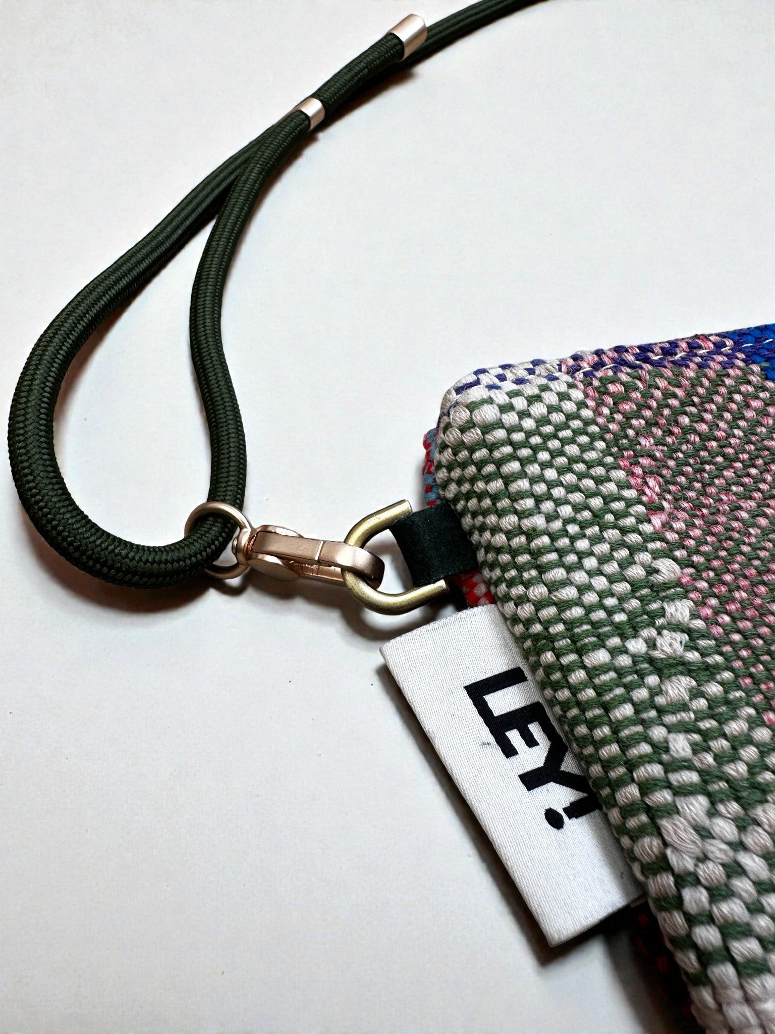 Eine Nahaufnahme eines grünen gewebten Schlüsselbands, das an der LEY! Tasche - OCULIA CLUTCH von LEY! aus Vintage-Stoff mit einem Metallverschluss. Ein weißes Schild mit der Aufschrift "LEY!" ist neben der Tasche teilweise sichtbar.