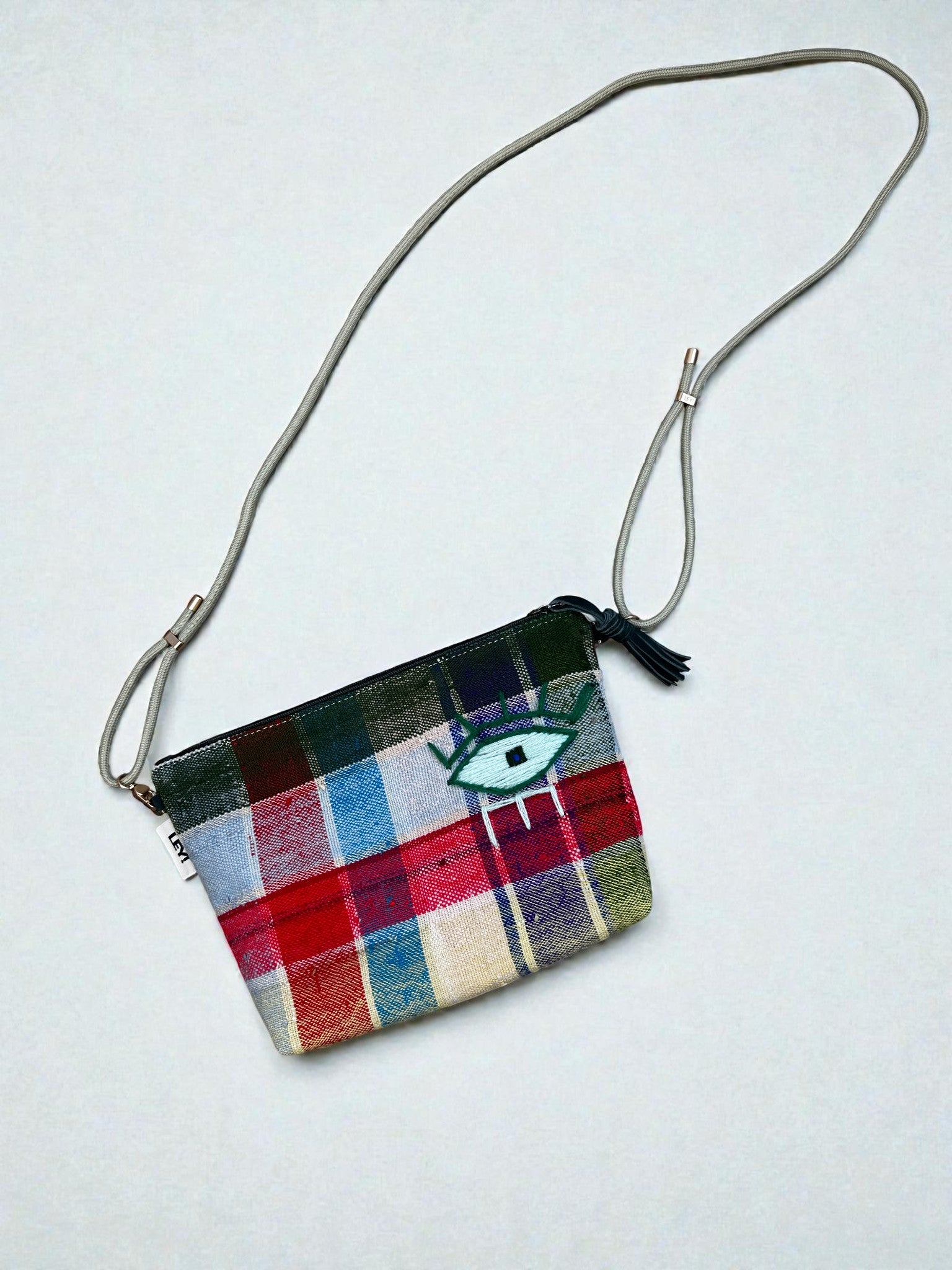 Die LEY! Tasche - OCULIA CLUTCH von LEY! ist eine kleine, handgefertigte Umhängetasche aus Vintage-Stoff mit Karomuster in Rot, Blau, Grün und Creme. Sie hat einen Oculia Clutch Aufnäher, Reißverschluss, Quaste und verstellbaren Träger.