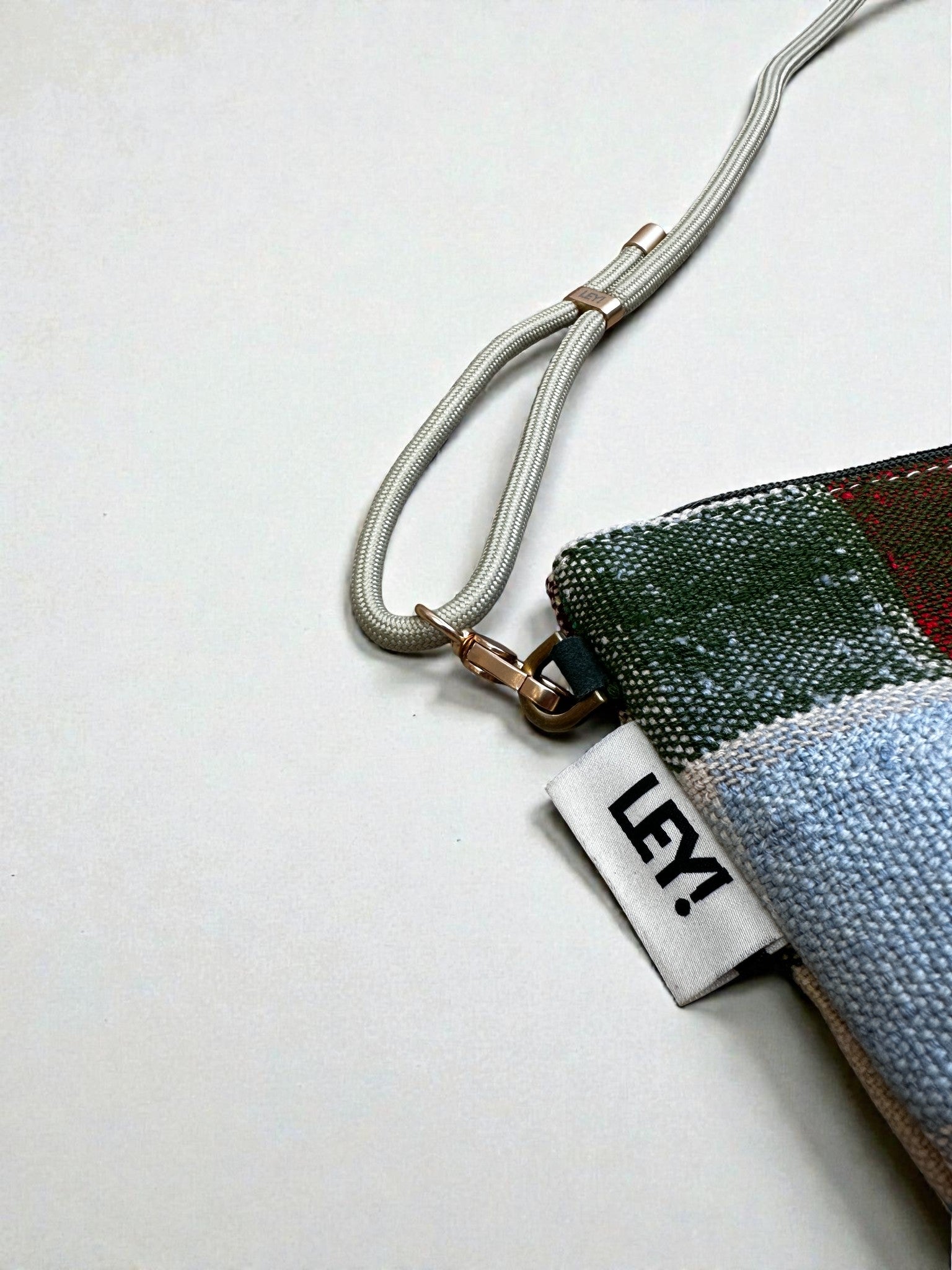Eine Nahaufnahme der LEY! Tasche - OCULIA CLUTCH von LEY!: eine kompakte Tasche mit lebhaftem Muster, einem weißen "LEY!"-Anhänger und einem beigen Armband mit goldener Schließe, dargestellt auf einer schlichten weißen Fläche.
