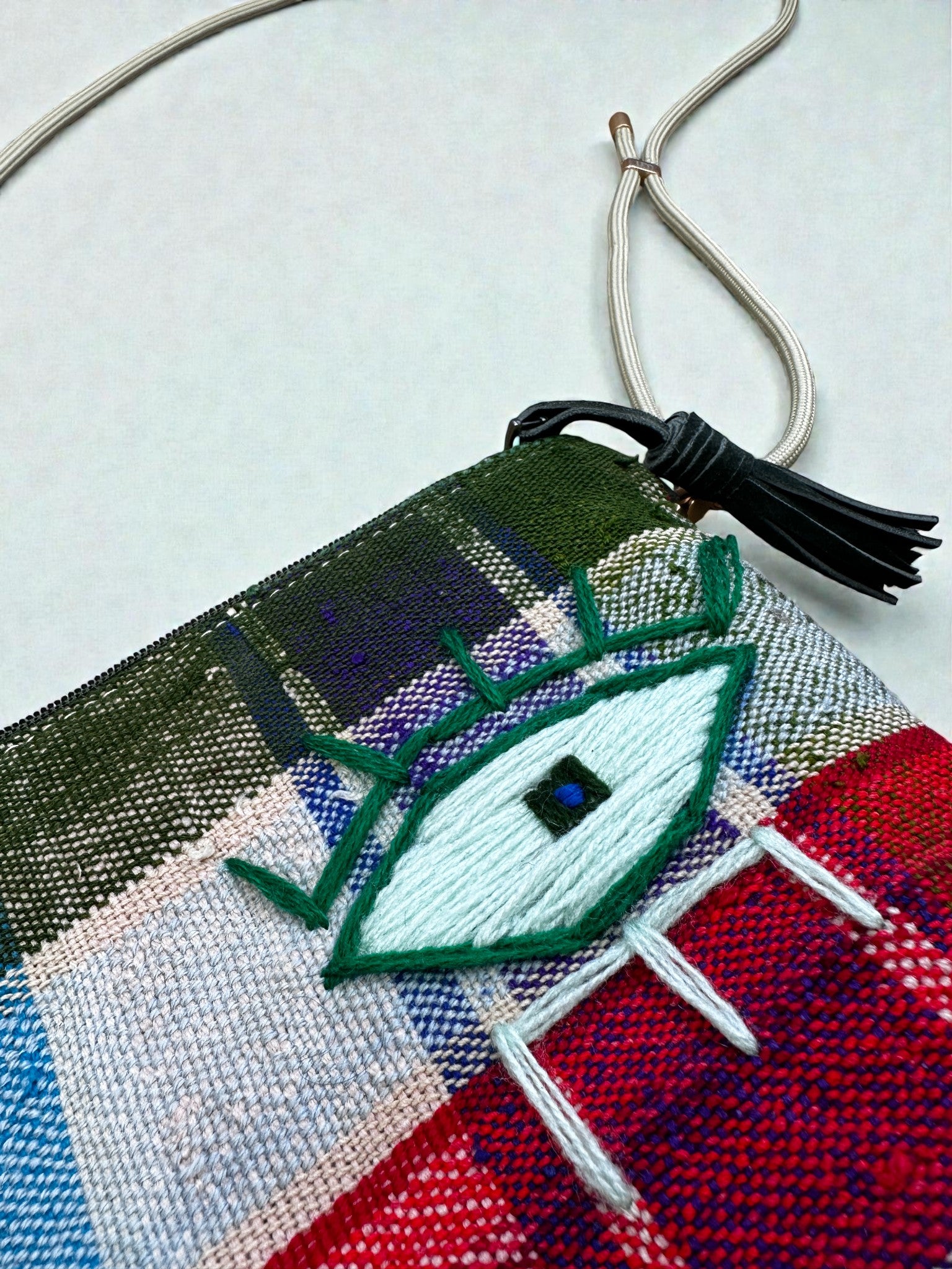 Die LEY! Tasche - OCULIA CLUTCH von LEY! ist eine farbenfrohe, handgefertigte Umhängetasche mit schwarzer Quaste und Kordelzug, die ein gesticktes grünes und weißes Auge mit blauen Details in grün, blau, rot und beige für einen lebendigen Look aufweist.