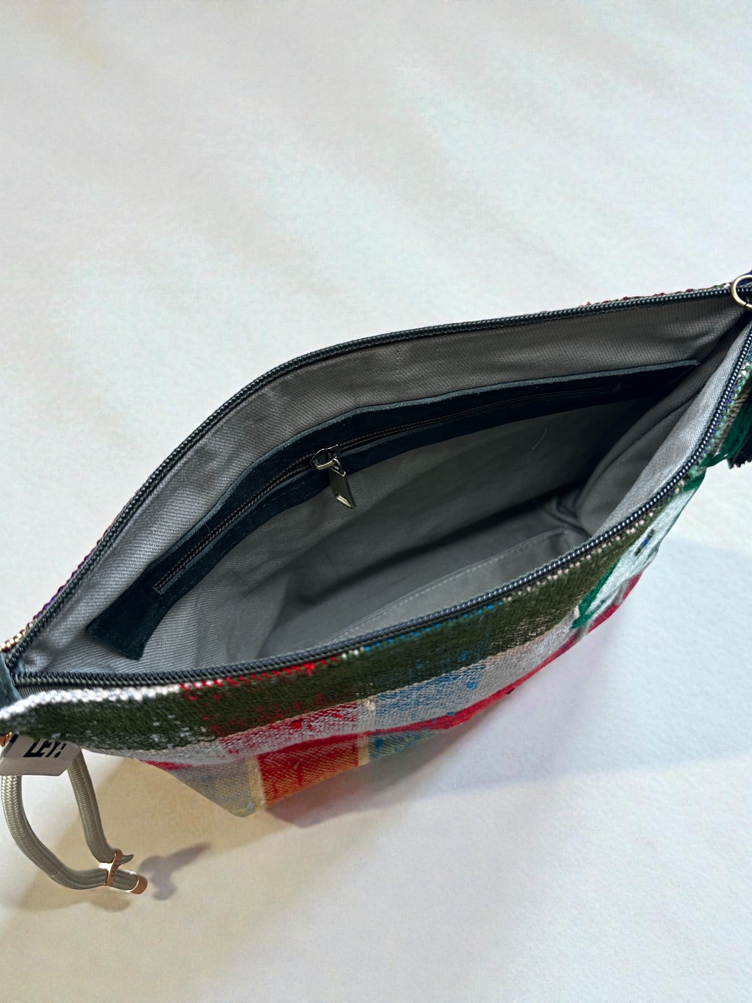 Ein Foto einer geöffneten LEY! Tasche - OCULIA CLUTCH von LEY! und zeigt das graue Innere mit einer Innentasche mit Reißverschluss. Die kompakte Clutch ist mit einem lebhaften, mehrfarbigen Karomuster versehen und steht auf einer hellen Oberfläche.