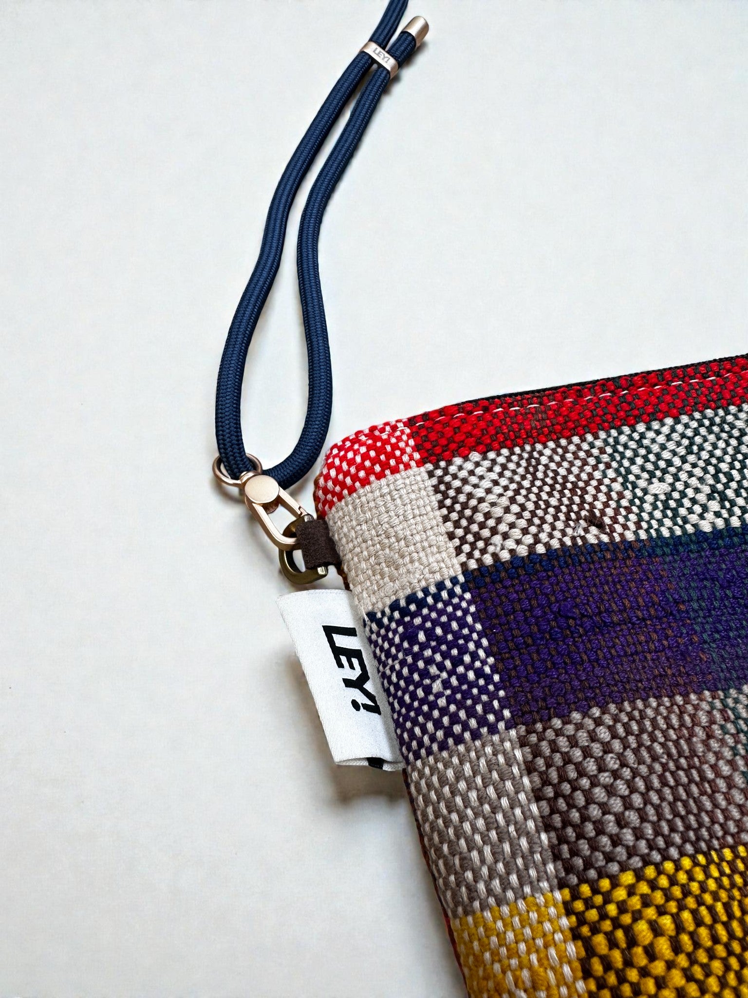 Eine Nahaufnahme der LEY! Tasche - OCULIA CLUTCH von LEY!: eine handgefertigte Umhängetasche mit rot, grau, lila, gelb und grün kariertem Vintage-Stoff, marineblauem Riemen und weißem "LEY!"-Anhänger.