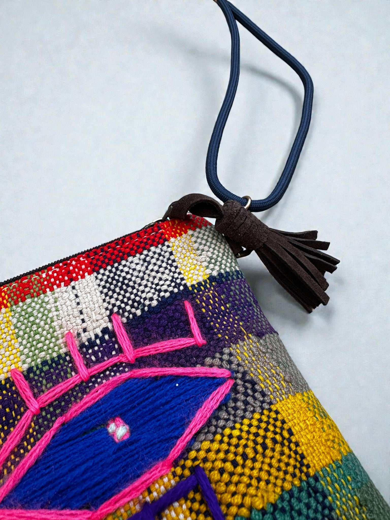 Eine Nahaufnahme der LEY! Tasche - OCULIA CLUTCH von LEY!, einer farbenfrohen, gewebten Tasche mit Quasten-Reißverschluss und blau-rosa gesticktem Augenmotiv auf Vintage-Stoff, vor einem karierten Hintergrund.