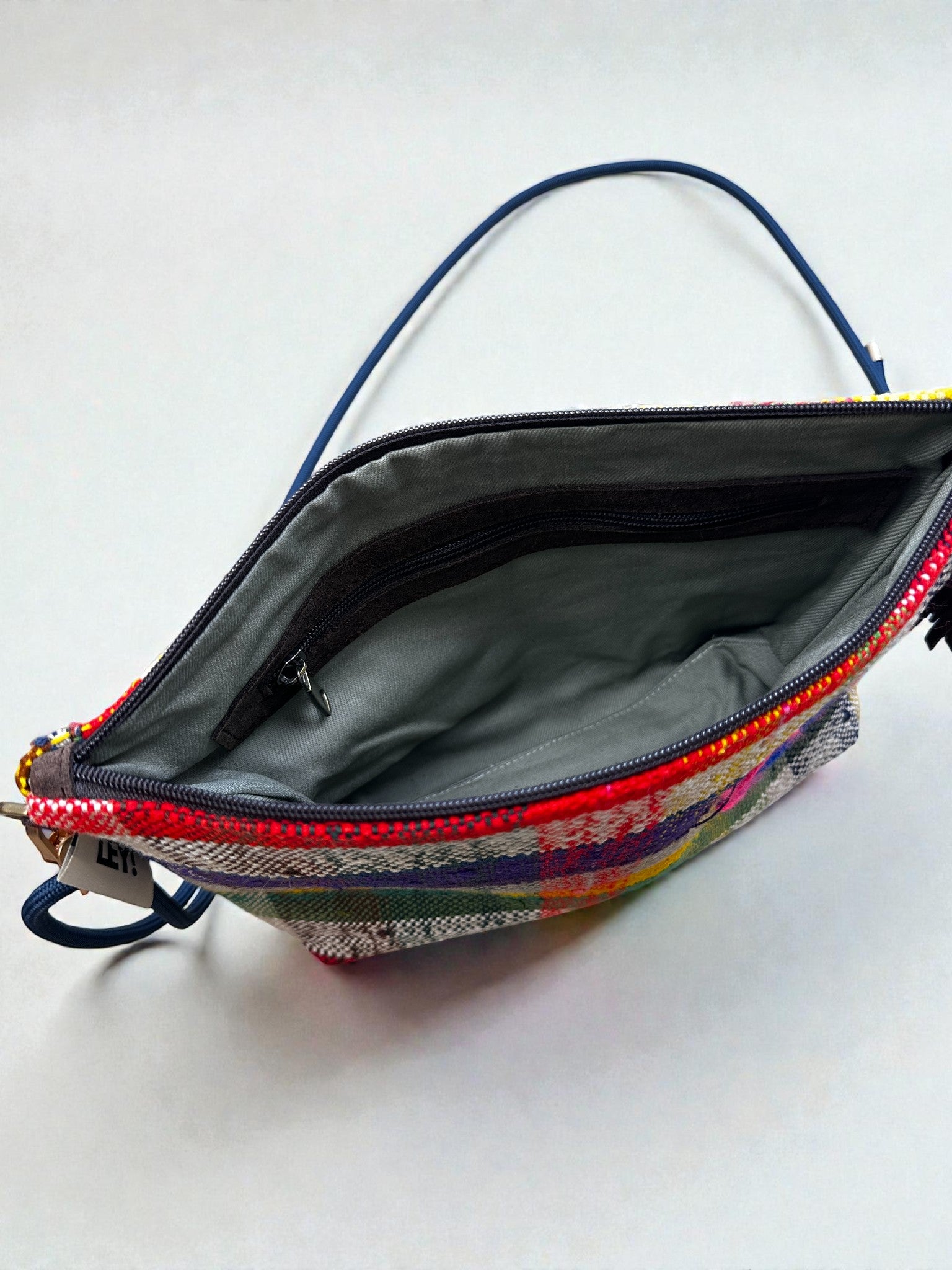 Die LEY! Tasche - OCULIA CLUTCH von LEY! ist eine farbenfrohe, karierte Reißverschlusstasche mit einer blauen Handschlaufe, die geöffnet gezeigt wird, um das graue Innenfutter, die Innentasche mit Reißverschluss und ein kleines weißes Etikett am Reißverschluss zu zeigen.
