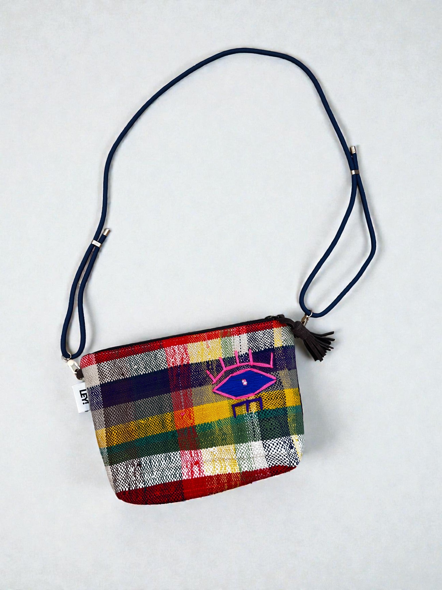 Die LEY! Tasche - OCULIA CLUTCH von LEY! ist eine kleine, handgefertigte Crossbody-Tasche mit einem mehrfarbigen Karomuster, einem verstellbaren, marineblauen Riemen, einem Reißverschluss und einem rosa-blauen, gestickten Augenmotiv mit schwarzer Quaste auf der Vorderseite.