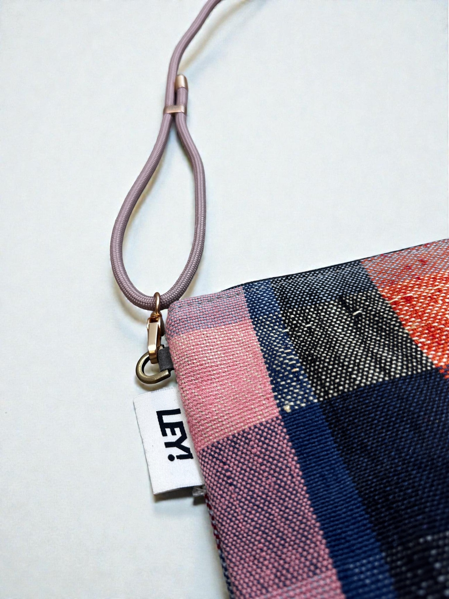 Eine Nahaufnahme der LEY! Tasche - OCULIA CLUTCH von LEY! aus Vintage-Stoff in einem rosa, blau, schwarz und rot gewebten Karomuster mit weißem "LEY!"-Anhänger und einem rosa Riemen, der über einen Metallring befestigt ist.