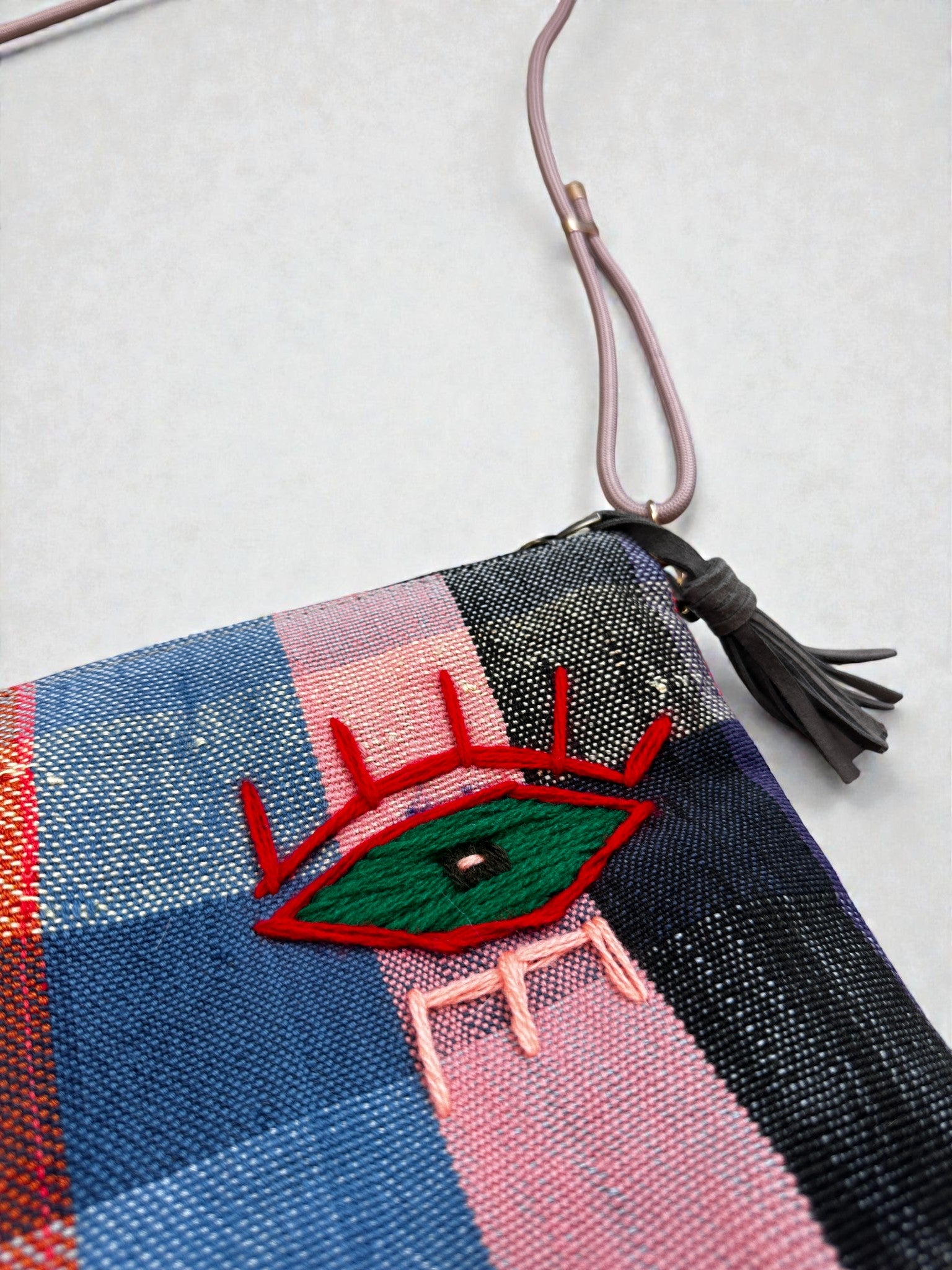Eine Nahaufnahme der LEY! Tasche - OCULIA CLUTCH von LEY! zeigt das farbenfrohe Karomuster, den rosa Riemen, die graue Quaste und ein gesticktes grünes Auge mit roten Wimpern und rosa Linien.