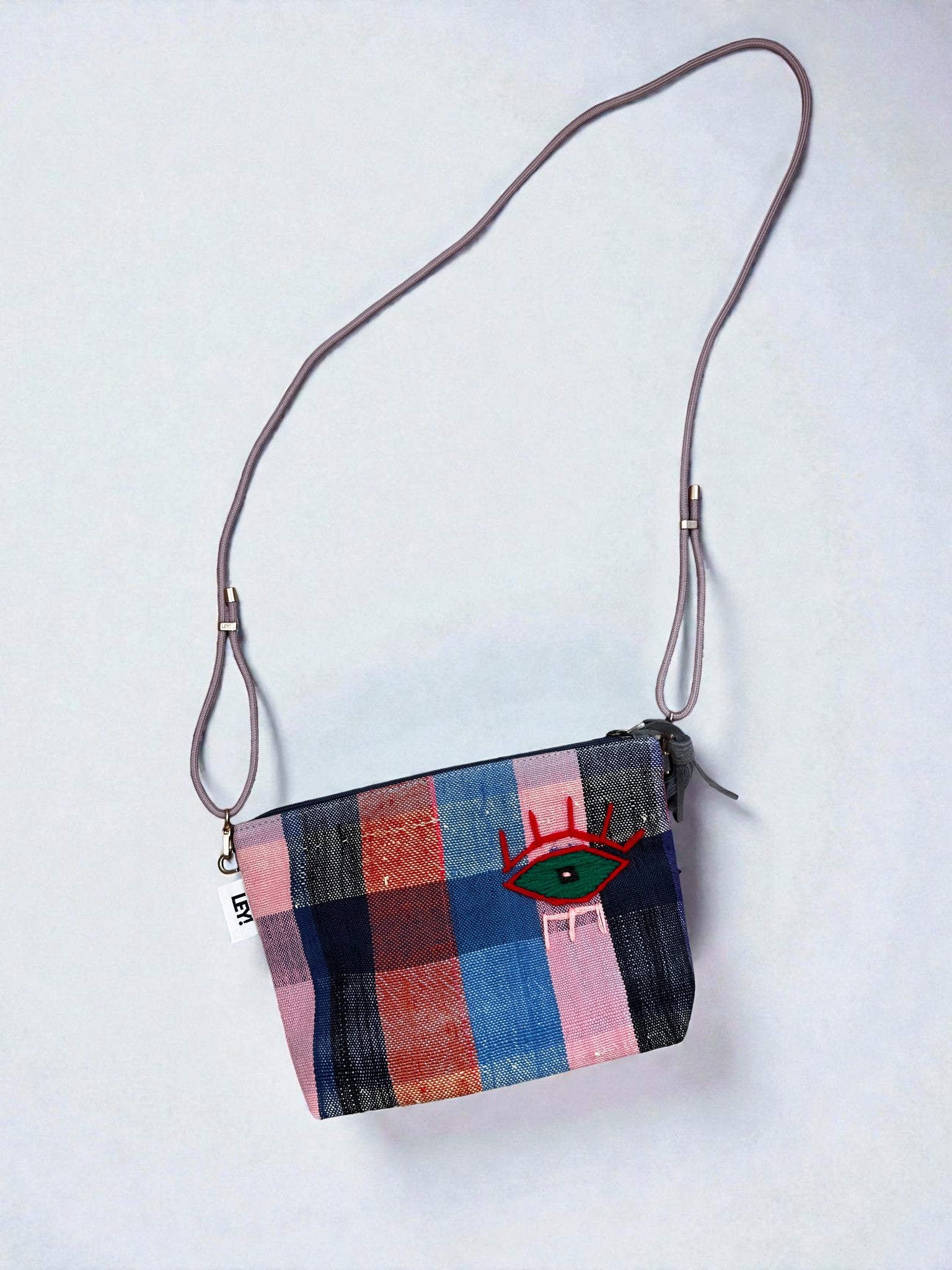 Die LEY! Tasche - OCULIA CLUTCH von LEY! ist eine kleine, handgefertigte Crossbody-Tasche mit einem langen, verstellbaren Riemen, einem Karomuster in Pink, Blau, Rot und Grau und einem gestickten Auge mit roten Wimpern sowie einem weißen Seitenanhänger.
