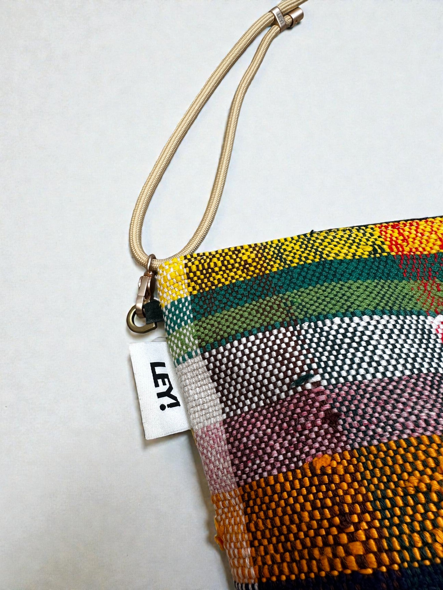 Die LEY! Tasche - OCULIA CLUTCH von LEY! ist eine farbenfrohe, gewebte Tasche mit beiger Handschlaufe und weißem "LEY!"-Anhänger auf weißem Grund - ideal als kompakte Schultertasche für den stilvollen Transport von Essentials.