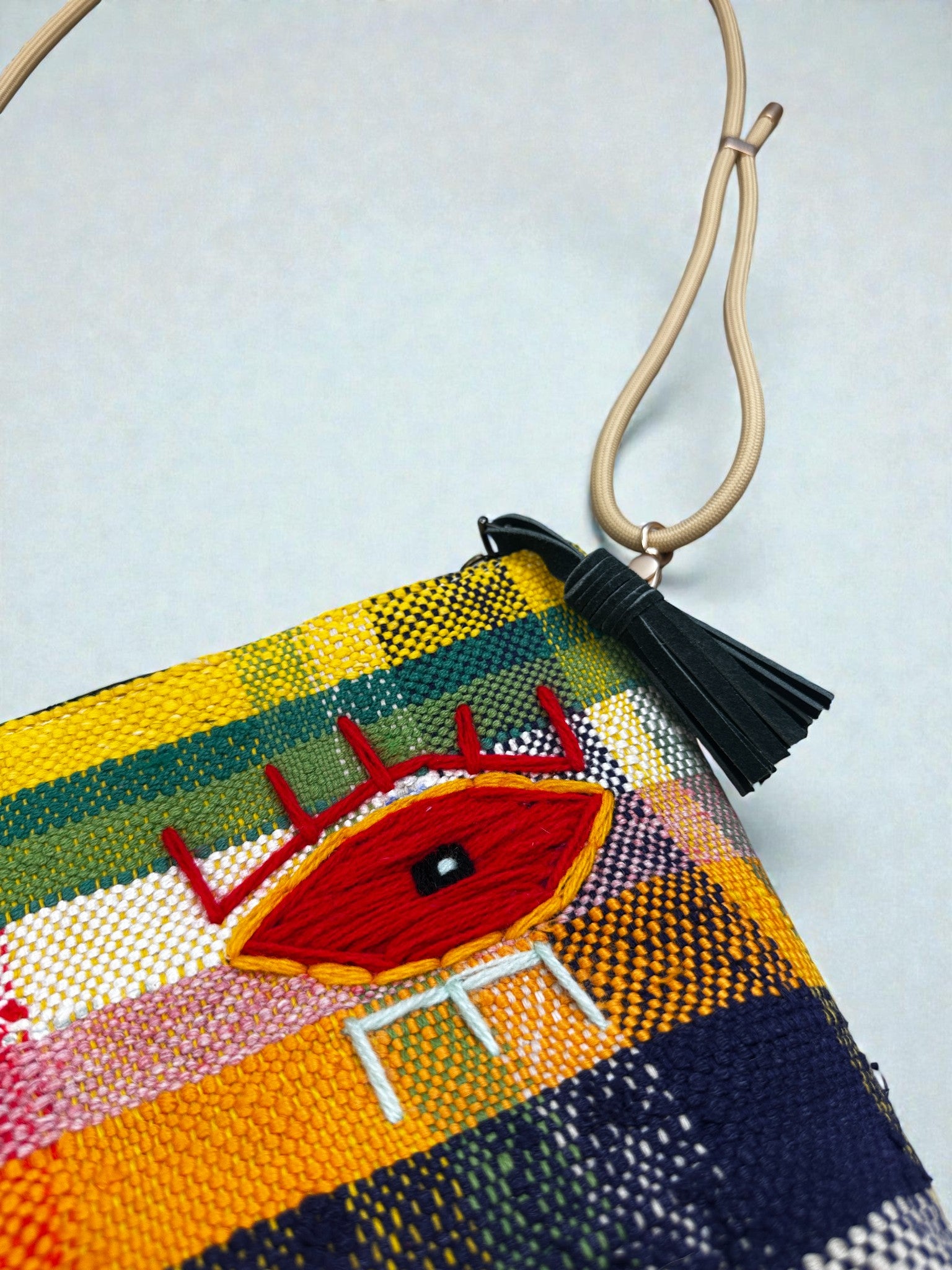 Die LEY! Tasche - OCULIA CLUTCH von LEY! ist eine farbenfrohe, gewebte Clutch mit einer roten, gestickten Öse, einer schwarzen Quaste und einem einzigartigen, hellbraunen Riemen, der vor einem hellen Hintergrund gezeigt wird.