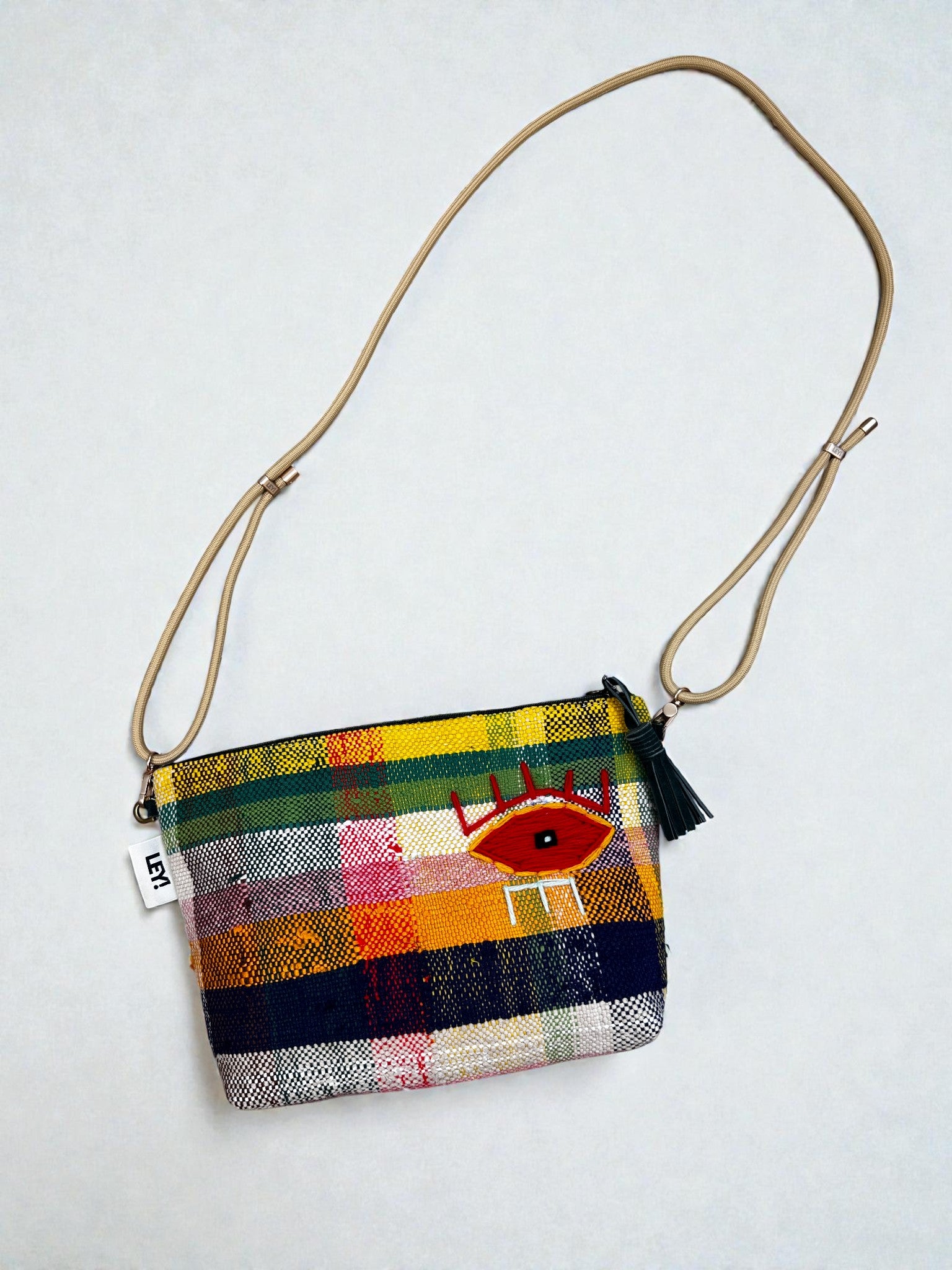 Die LEY! Tasche - OCULIA CLUTCH von LEY! ist eine handgefertigte Umhängetasche mit farbenfrohem Karomuster, braunem verstellbaren Riemen, schwarzer Quaste, gesticktem roten Auge mit Kronendetails und einem "LFT"-Anhänger - ideal als Festivaltasche.