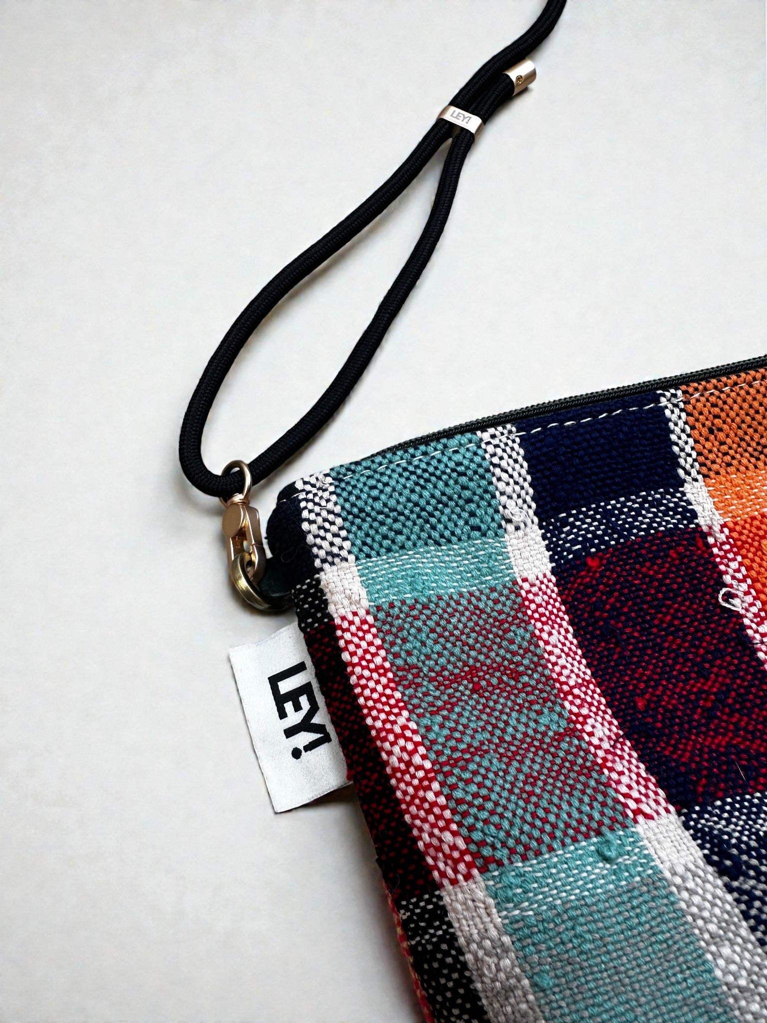 Eine Nahaufnahme der LEY! Tasche - OCULIA CLUTCH, einer lebhaft karierten Reißverschlusstasche mit schwarzer Handschlaufe und weißem "LEY!"-Anhänger - handgefertigt von LEY! für einen einzigartigen Look mit Vintage-Stoff.