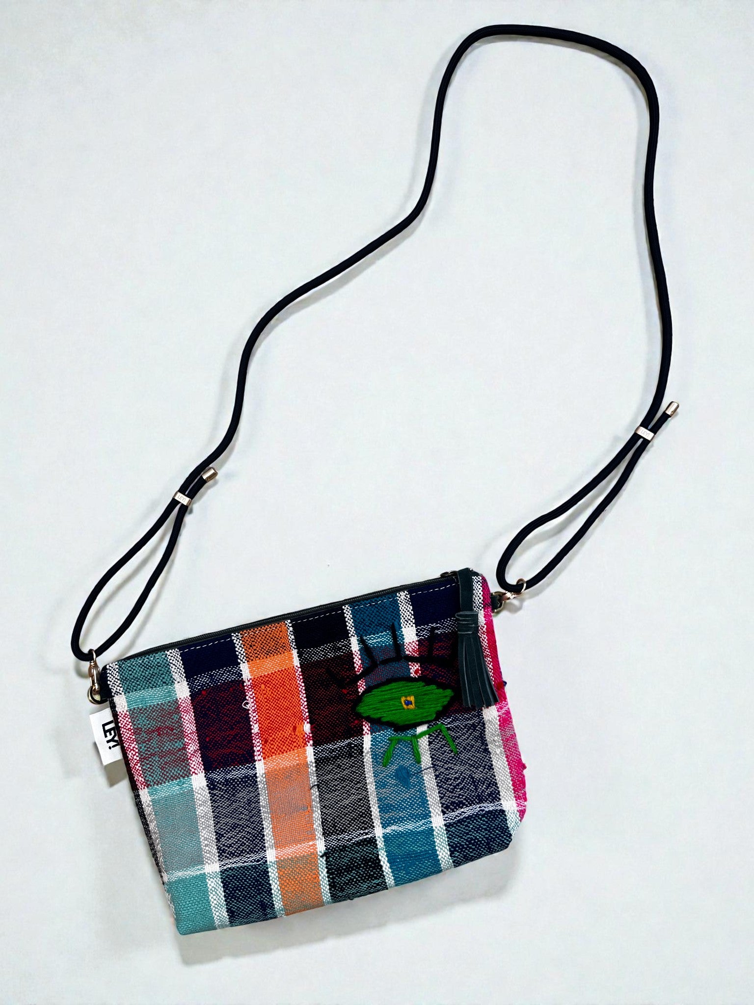 Die LEY! Tasche - OCULIA CLUTCH von LEY! ist eine handgefertigte Umhängetasche aus Vintage-Stoff im mehrfarbigen Plaid-Muster mit langem, verstellbarem schwarzem Gurt und grün gesticktem Oculia-Augenmotiv vorn.