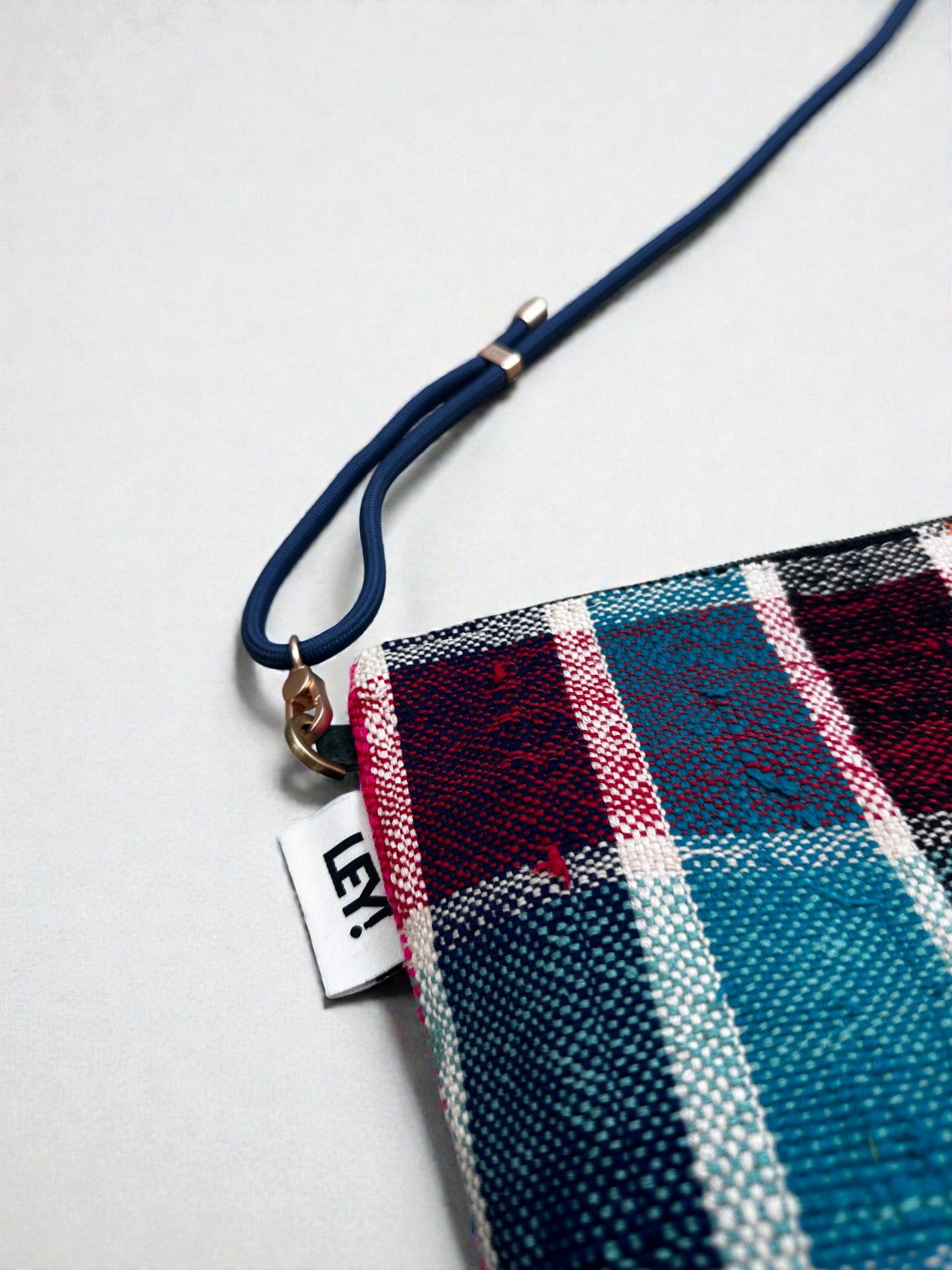 Eine Nahaufnahme der LEY! Tasche - OCULIA CLUTCH, einer kleinen karierten Stofftasche von LEY! mit blauen, roten und weißen Streifen, einer blauen Handschlaufe und einem weißen "LEY!"-Anhänger, die einen charmanten Vintage-Look auf hellem Grund bietet.
