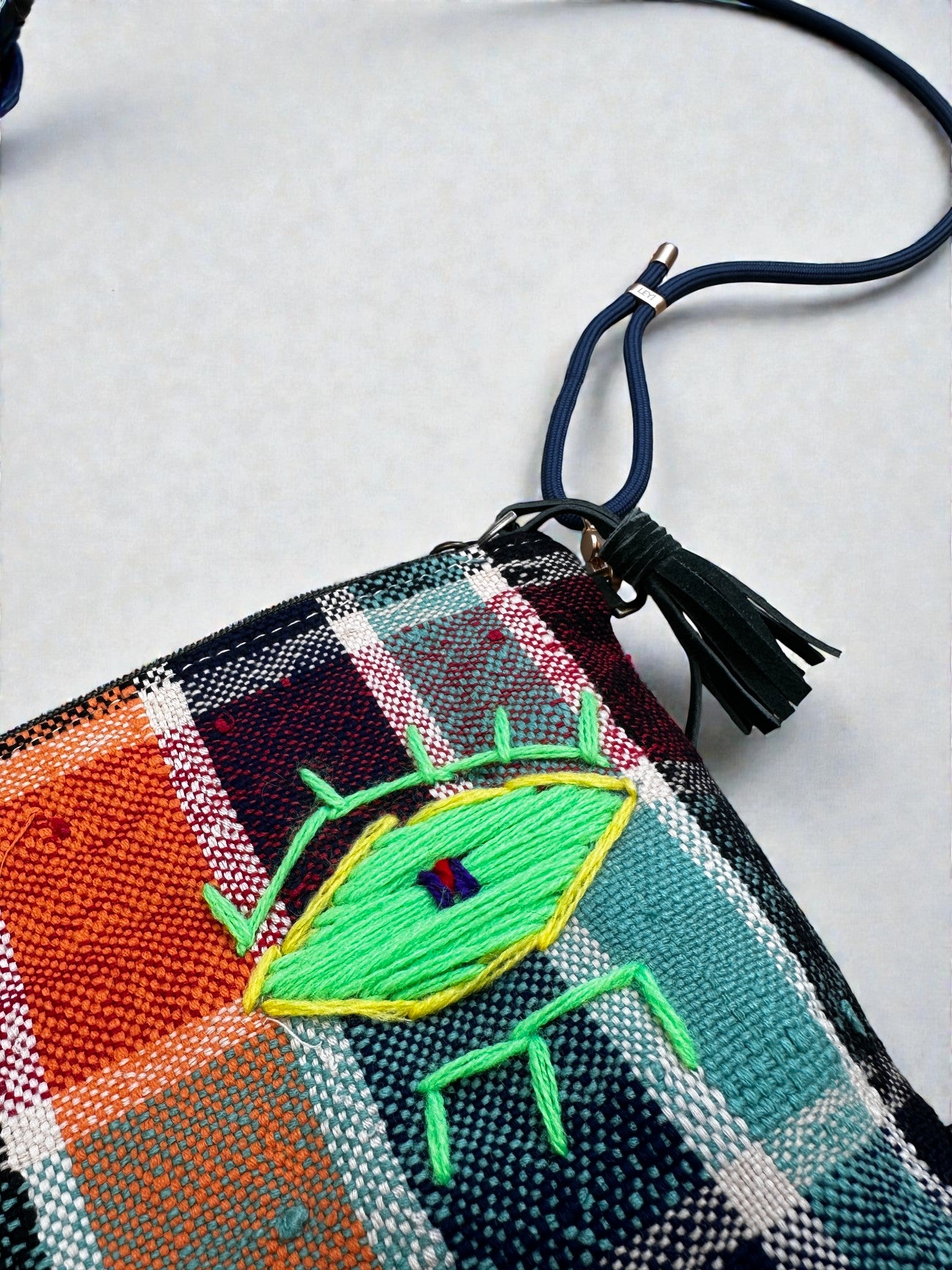 Die LEY! Tasche - OCULIA CLUTCH von LEY! ist eine handgefertigte Umhängetasche mit blauem Riemen, Quaste und gesticktem gelb-grünen Auge auf orange-, teal- und navy-kariertem Vintage-Stoff.