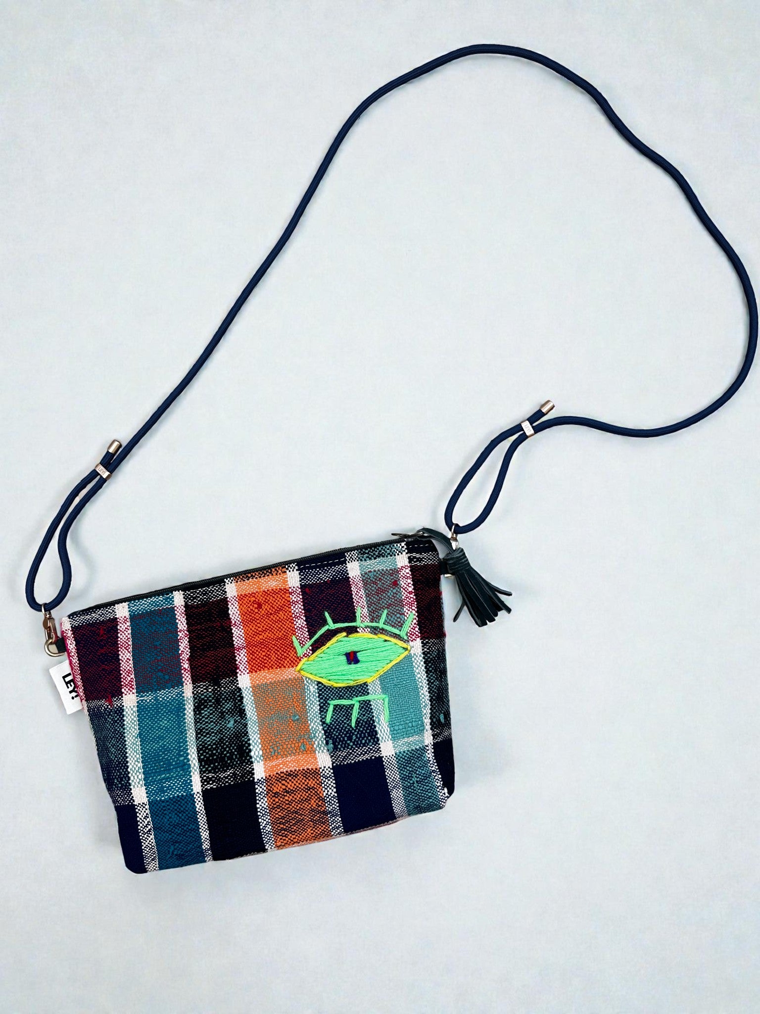 Die LEY! Tasche - OCULIA CLUTCH von LEY! ist eine kleine, handgefertigte Crossbody-Tasche mit blauem Riemen, farbenfrohem Karomuster und gesticktem grünen Auge auf der Vorderseite. Sie hat einen Reißverschluss mit einer dunklen Quaste.