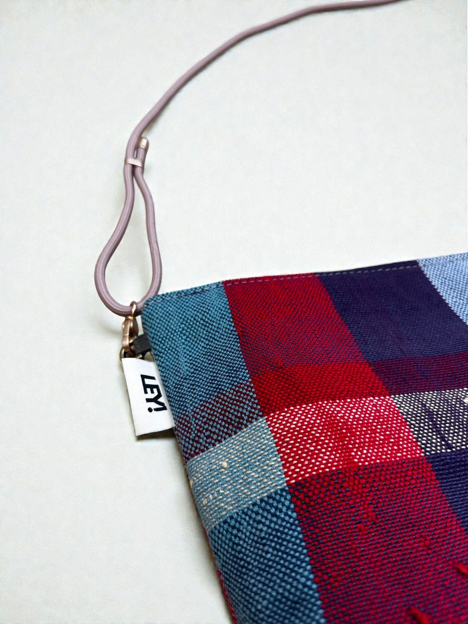 Eine Nahaufnahme der LEY! Tasche - OCULIA CLUTCH von LEY!, einer handgefertigten Umhängetasche aus rot, blau und lila kariertem Vintage-Stoff mit einem rosa Riemen und einem kleinen weißen "LEY!"-Anhänger auf hellem Hintergrund.