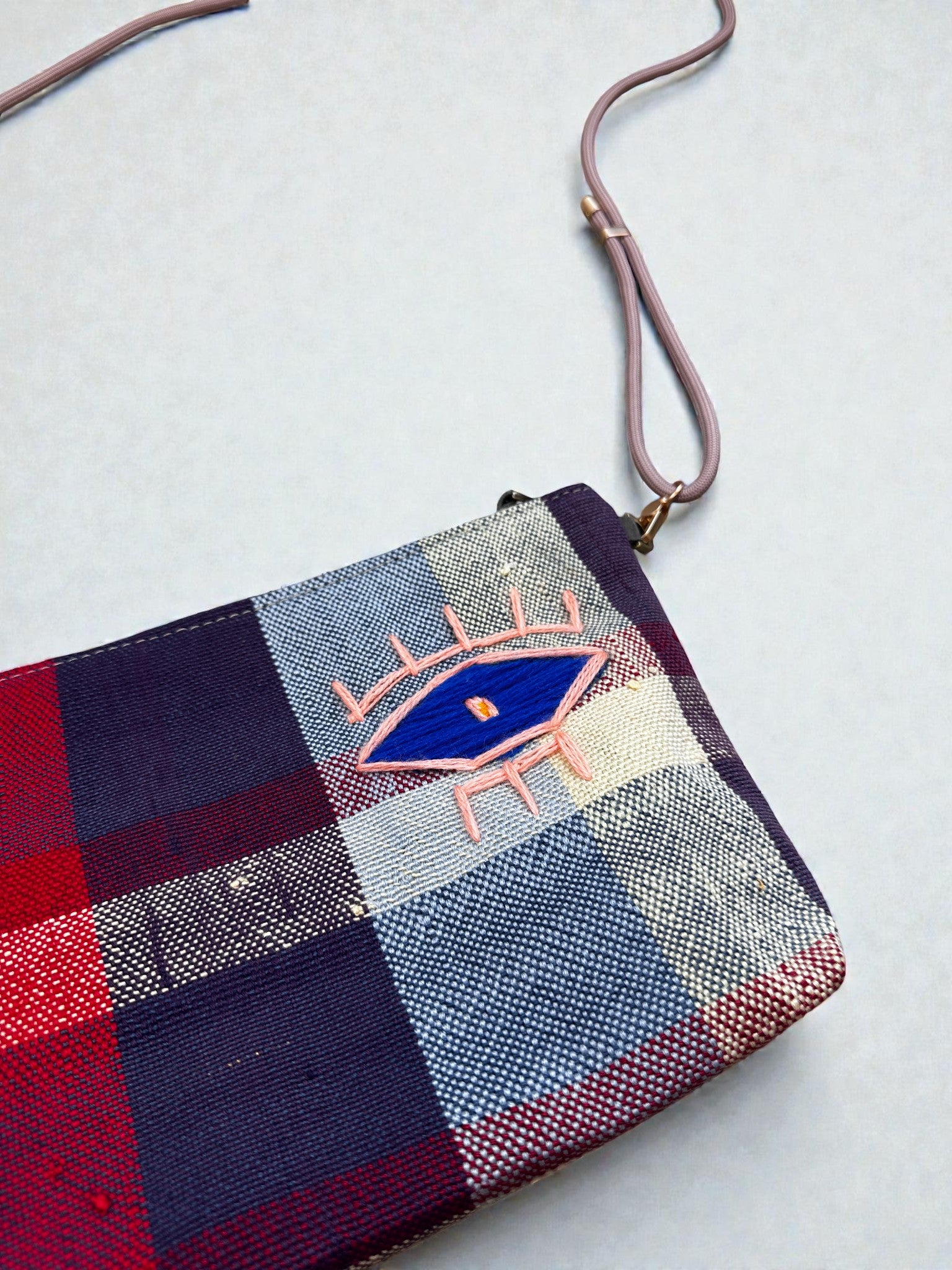 Die LEY! Tasche - OCULIA CLUTCH von LEY! ist eine karierte Geldbörse in blauem, rotem und weißem Vintage-Stoff mit einem gestickten Augenmotiv, rosa Wimpern und einem dünnen lavendelfarbenen Schulterriemen.