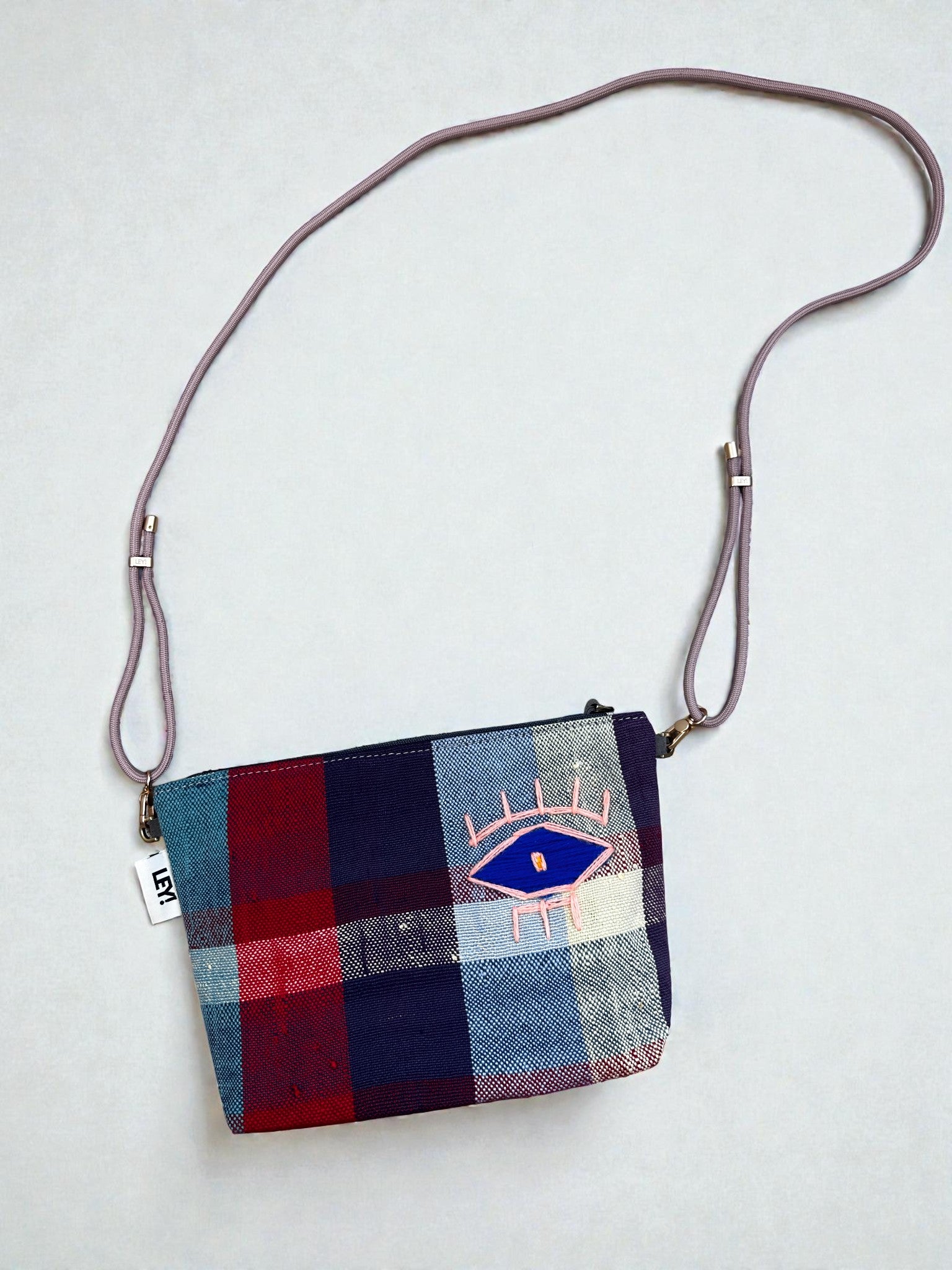 Die LEY! Tasche - OCULIA CLUTCH von LEY! ist eine kleine, rechteckige Umhängetasche aus Vintage-Stoff mit lila Träger, rot-blau-weiß kariertem Muster, gesticktem Auge vorne und "HEY"-Anhänger am Rand.