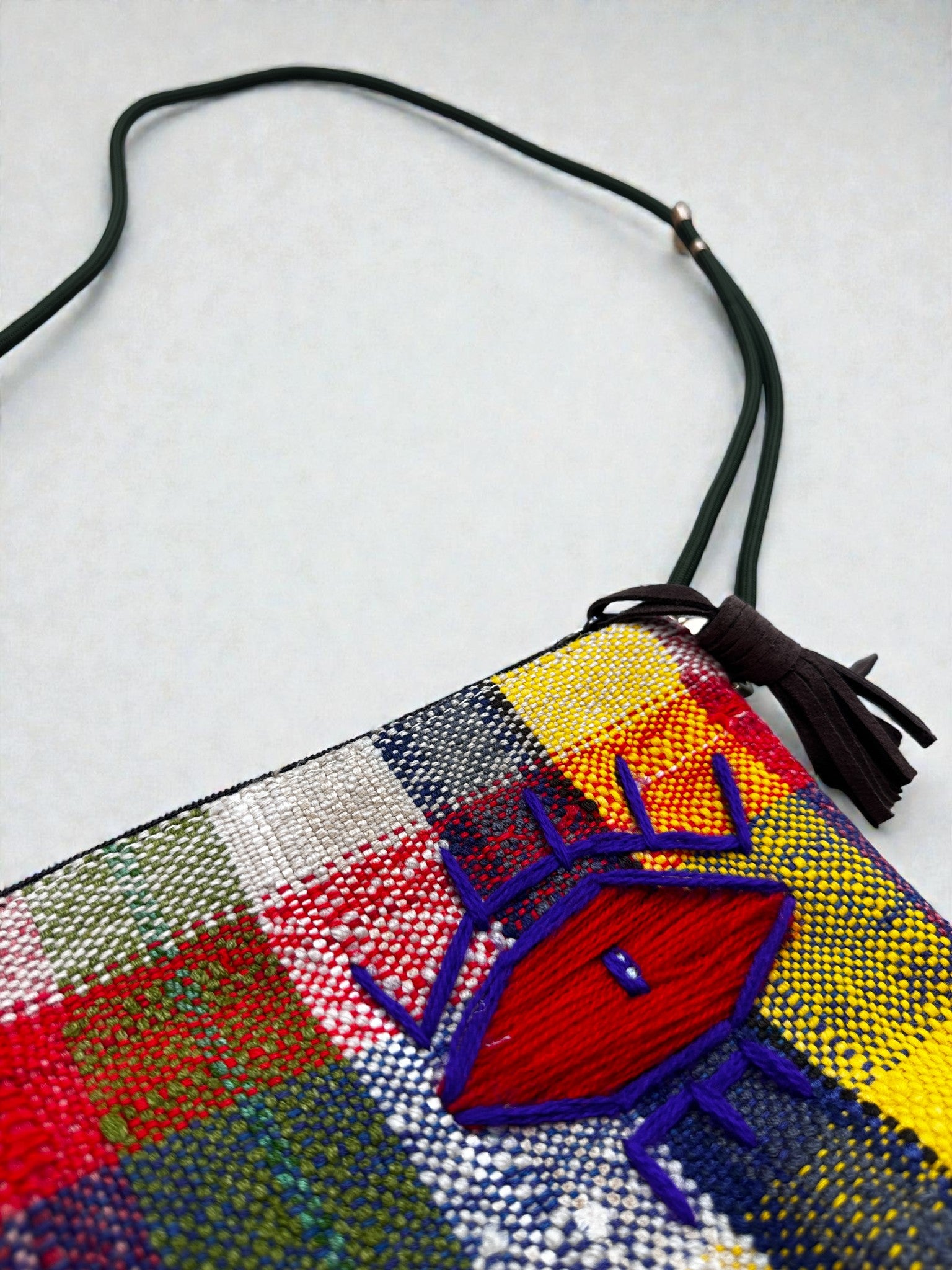 Die LEY! Tasche - OCULIA CLUTCH von LEY! ist eine farbenfrohe, karierte Crossbody-Tasche aus Stoff mit einem rot und lila gestickten Auge, einem grünen Riemen und einer braunen Quaste, die auf einem weißen Hintergrund abgebildet ist.