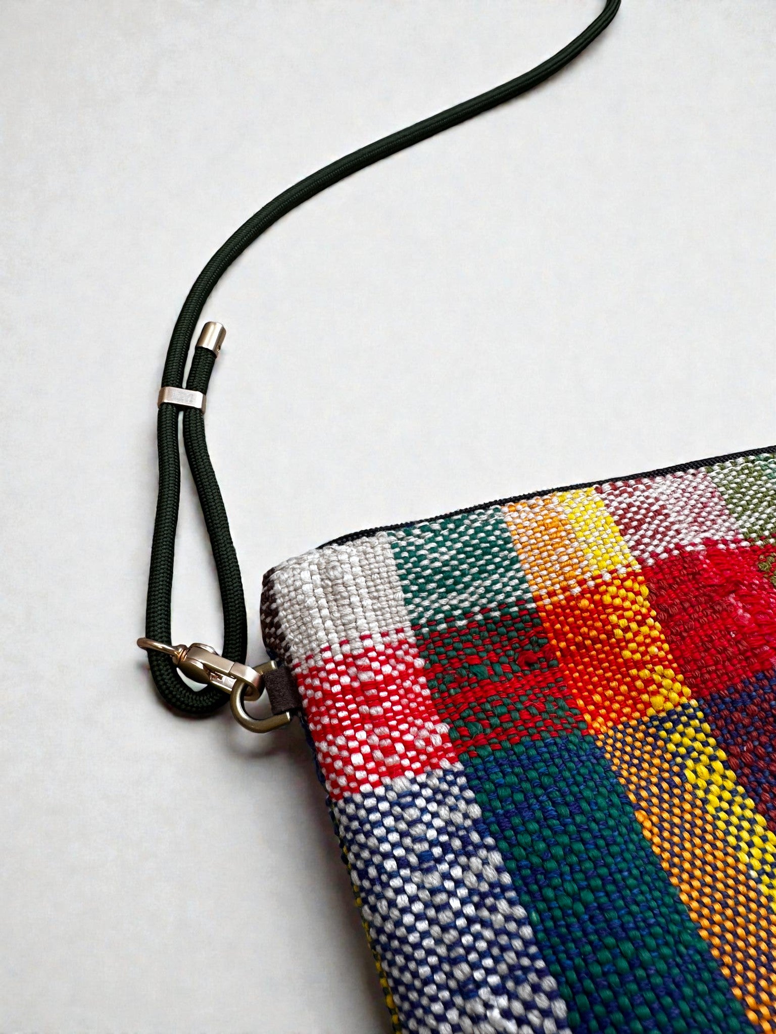 Eine Nahaufnahme der LEY! Tasche - OCULIA CLUTCH von LEY!, einer farbenfrohen Clutch mit einem rot, grün, blau, gelb und weiß karierten Muster. Sie hat einen dunkelgrünen Riemen mit Metallverschluss vor einem schlichten hellen Hintergrund.