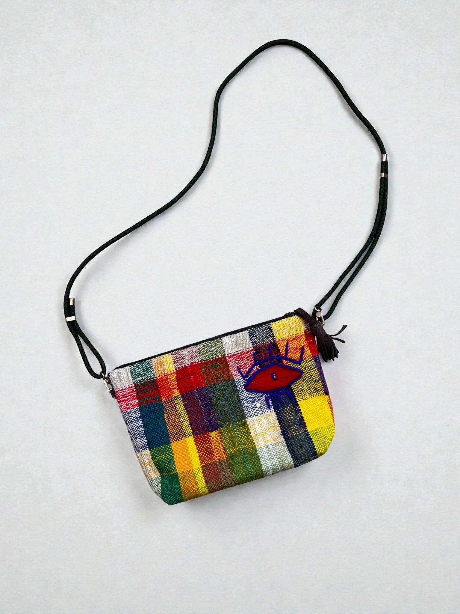 Die LEY! Tasche - OCULIA CLUTCH von LEY! ist eine kleine, handgefertigte Umhängetasche mit einem leuchtenden Karomuster in Gelb, Grün, Rot und Blau, einem gestickten blauen Auge und einem schwarzen, verstellbaren Riemen - perfekt für jeden Anlass.