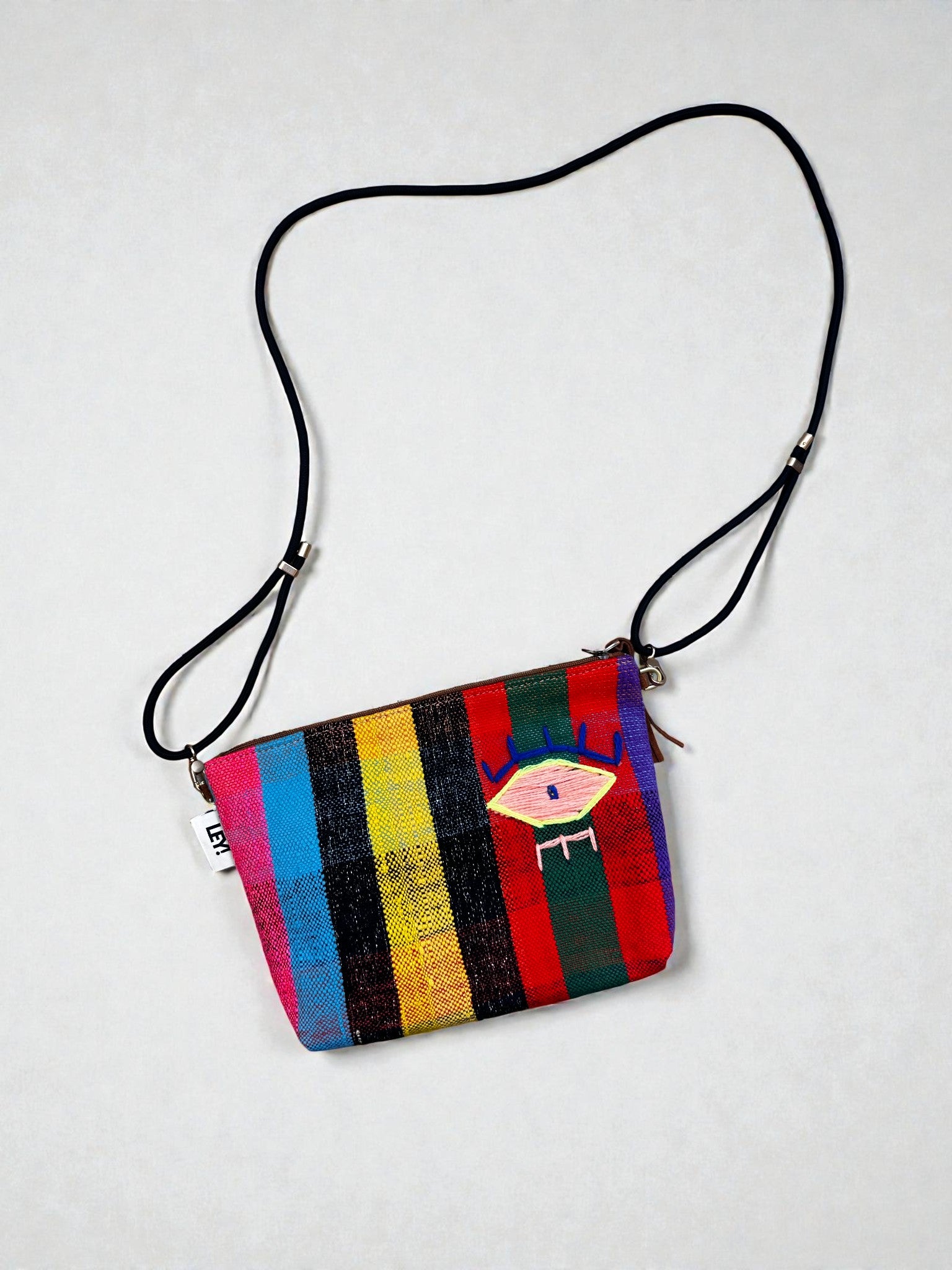 Die LEY! Tasche - OCULIA CLUTCH von LEY! ist eine kleine, farbenfrohe Umhängetasche mit vertikalen Regenbogenstreifen, einem schwarzen Riemen und einem gestickten Augenmotiv auf der Vorderseite, die aus einem einzigartigen Vintage-Stoff gefertigt ist.